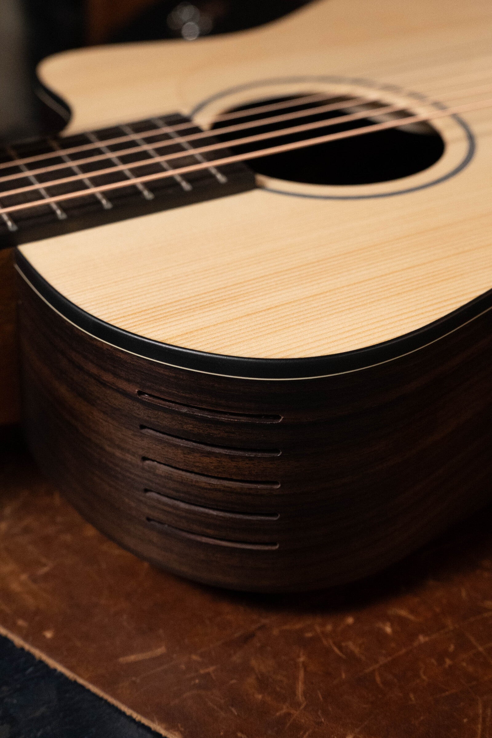 Close-up of the Furch Pioneer-ER VTC travel guitar, featuring an Engelmann Spruce top, Indian Rosewood sides, sound hole, strings, horizontal soundports with Boosters, and a VTC pickup. The cutaway body rests on a brown leather surface.