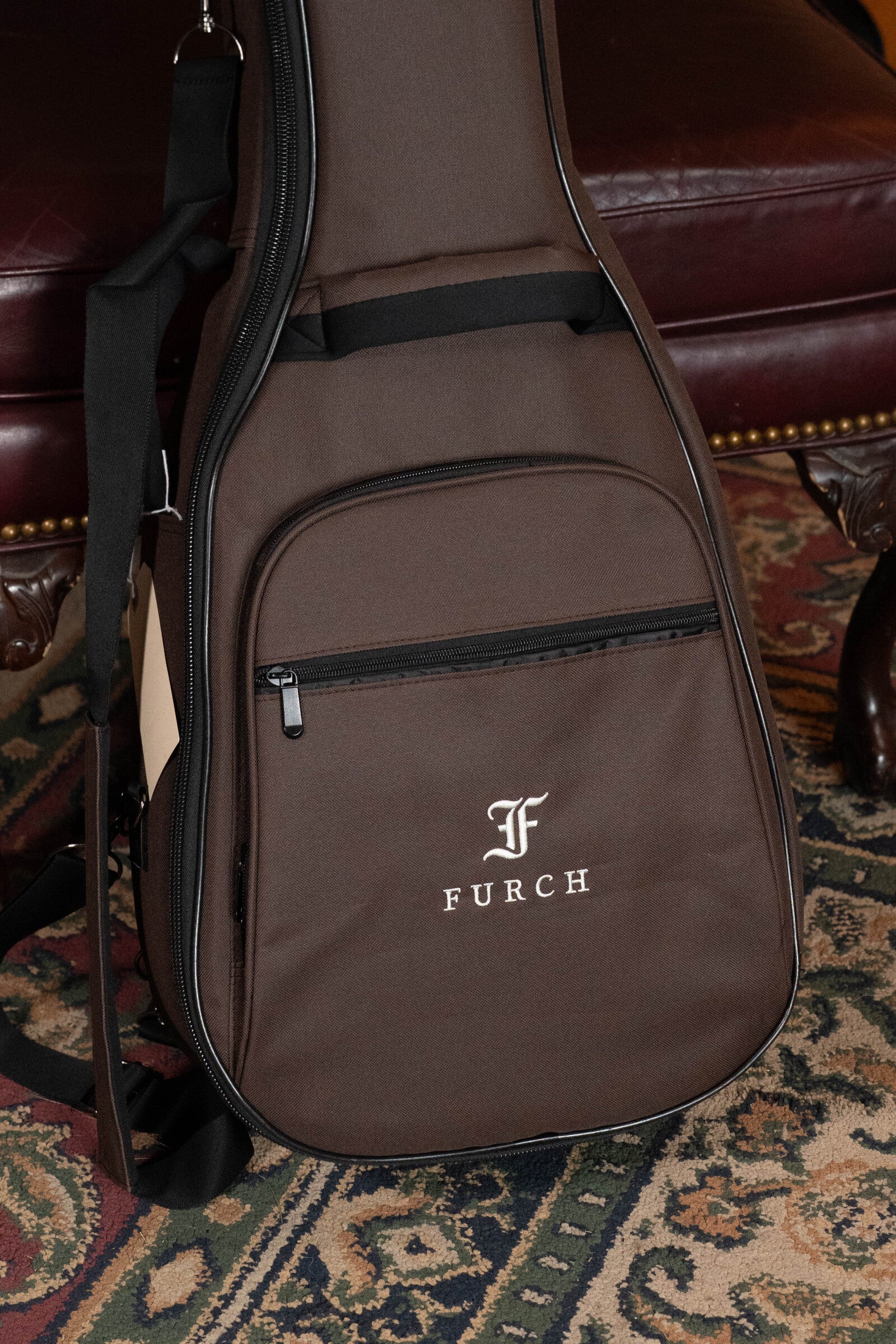 A Furch Pioneer-ER VTC Engelmann Spruce/Indian Rosewood Cutaway Travel Guitar with Soundport Boosters and VTC Pickup (#0808) in a brown Furch gig bag sits on a patterned rug next to a dark red leather chair with brass studs.