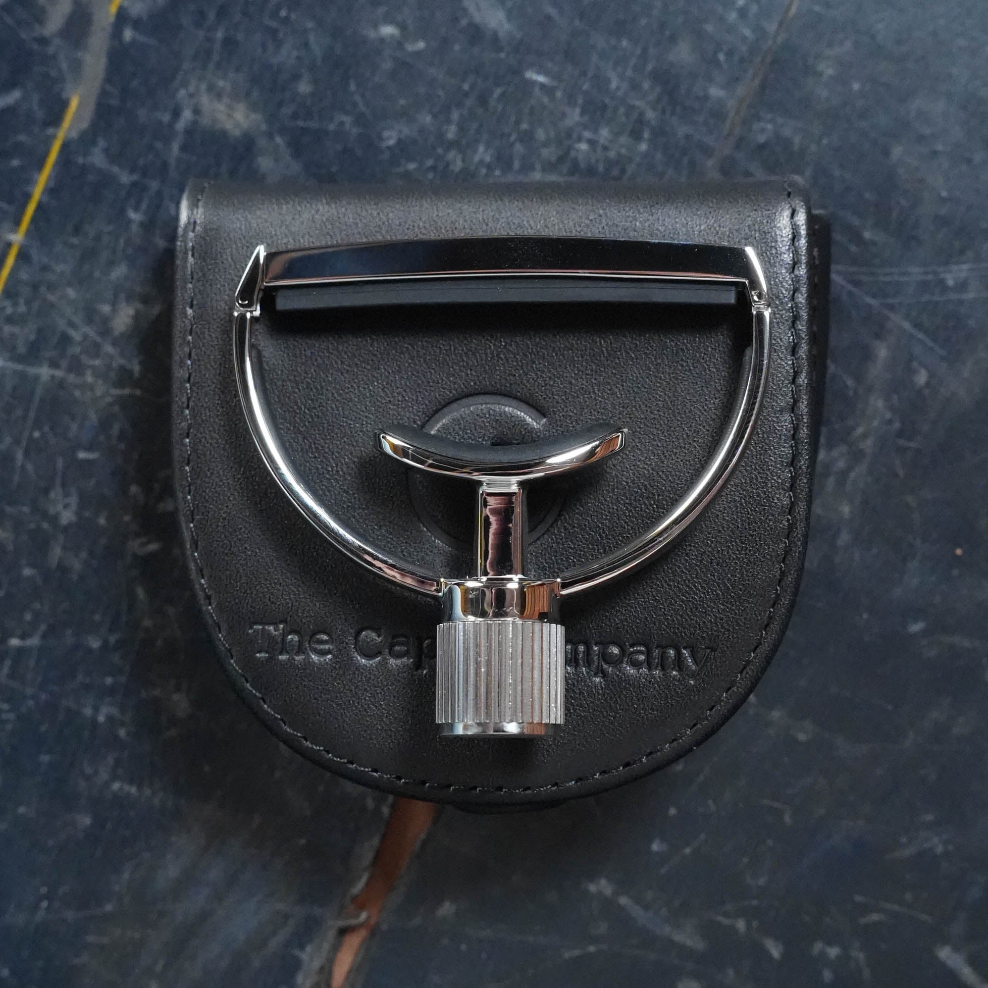 A gleaming G7th Heritage Guitar Capo S1 Wide with a knurled handle rests on a black leather pouch embossed with “The Acoustic Shoppe,” echoing the precision of their capos, atop a dark, scratched surface.