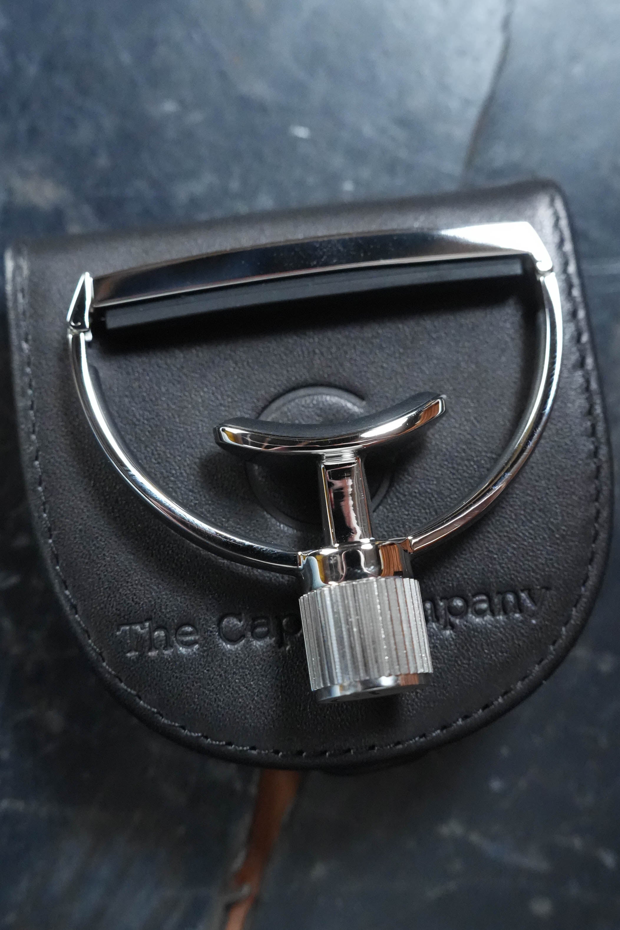 A shiny metal G7th Heritage Guitar Capo S1 Wide by The Acoustic Shoppe rests on a black leather case embossed with the brand, its knurled handle and design echoing the precision of their renowned yoke-style capo, on a dark textured surface.