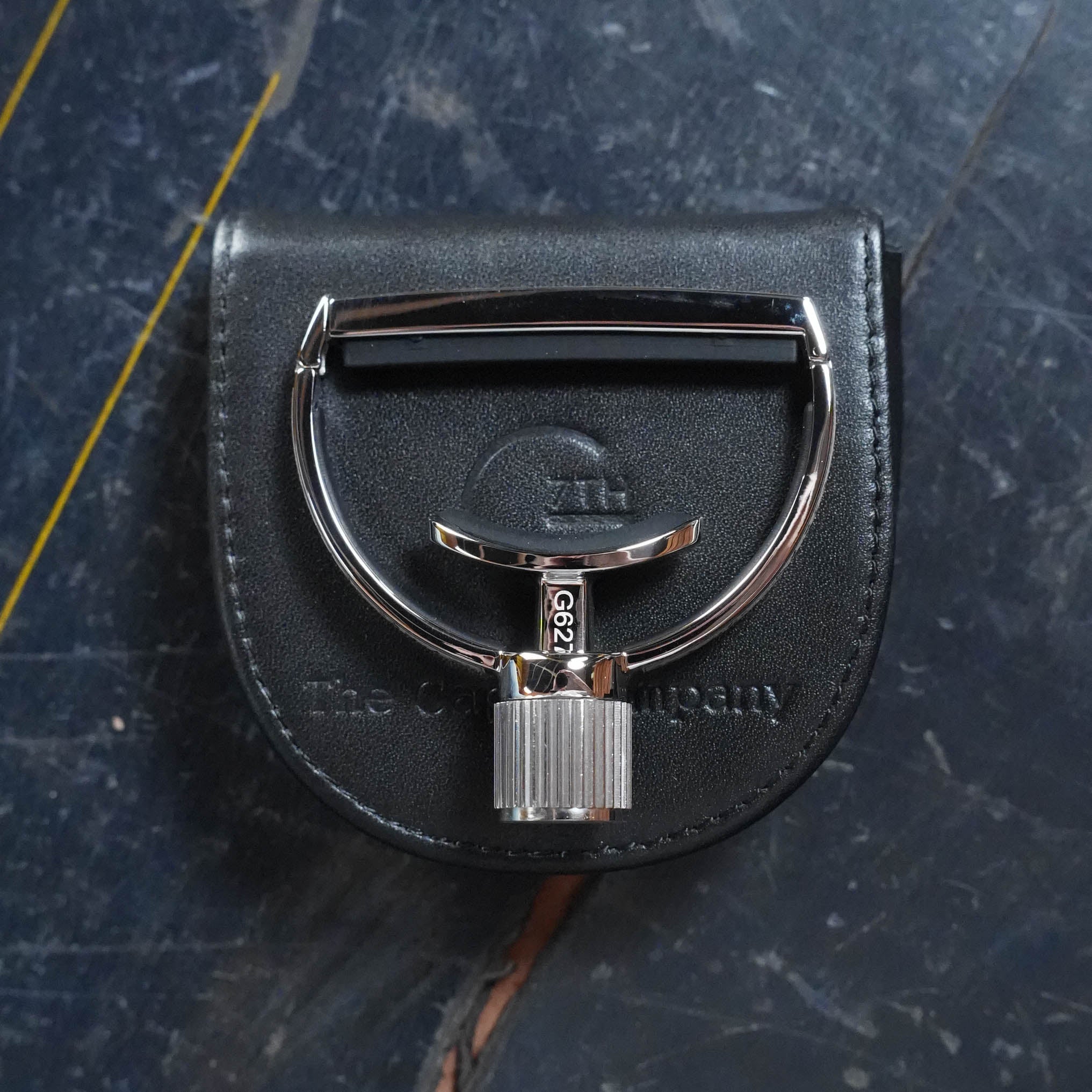 A shiny silver belt buckle with a unique design sits on a black leather case embossed with The Acoustic Shoppe, next to the G7th Heritage Guitar Capo S3 Standard Neck Width, all arranged on a marbled dark surface with yellow and brown streaks.