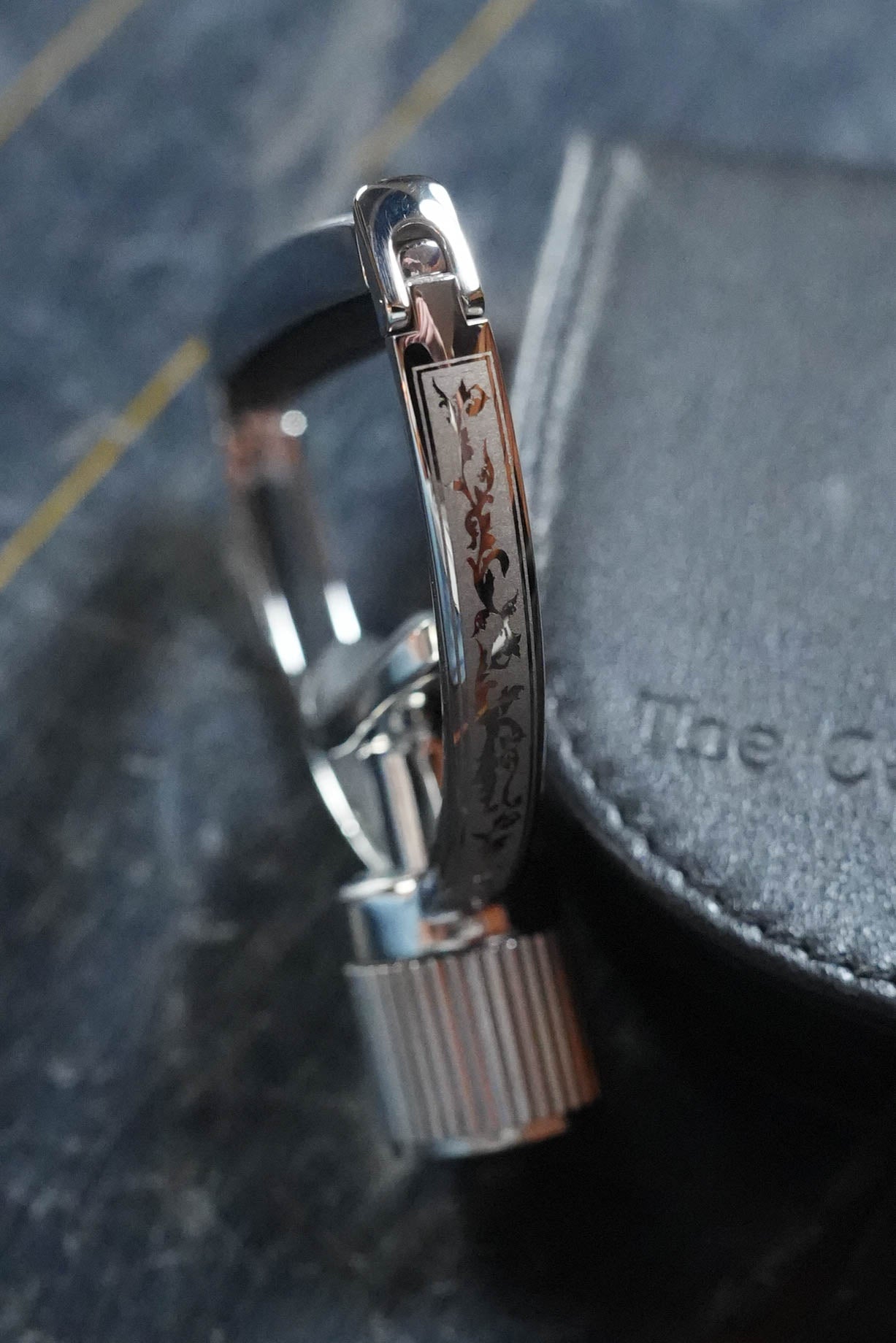 A close-up of a metallic watch clasp with Japanese engravings, beside a black leather case and a G7th Heritage Guitar Capo S3 Wide Engraved by The Acoustic Shoppe, featuring Adaptive Radius Technology on a dark textured surface.