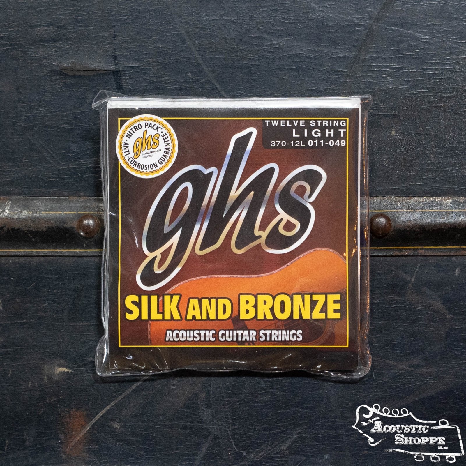 A package of GHS Strings Silk and Bronze 12 String Light (370-12L) acoustic guitar strings sits on a dark surface. The brown packaging with yellow and white text is ideal for any semi-acoustic twelve-string guitar.