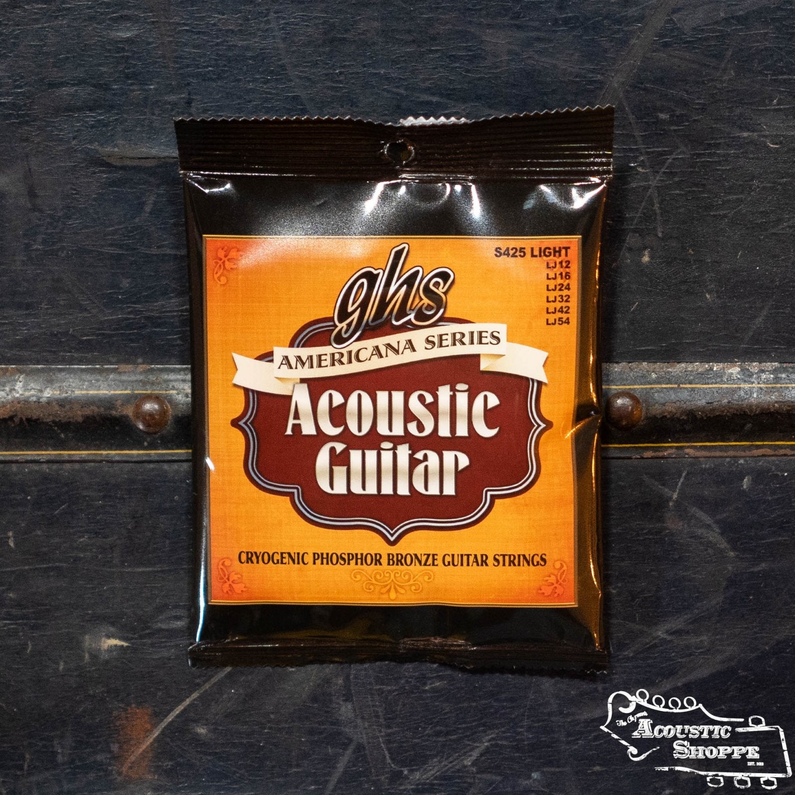 A pack of GHS S425 Americana Series Acoustic Guitar Strings - Light from GHS Strings sits upright on a dark surface, featuring an orange and brown label. The Acoustic Shoppe logo is visible in the corner.