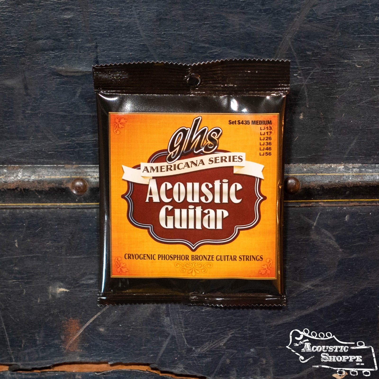 A pack of GHS Strings S435 Americana Guitar Medium strings, perfect for medium acoustic tones, is shown on a dark wooden surface beside “The Acoustic Shoppe” logo.
