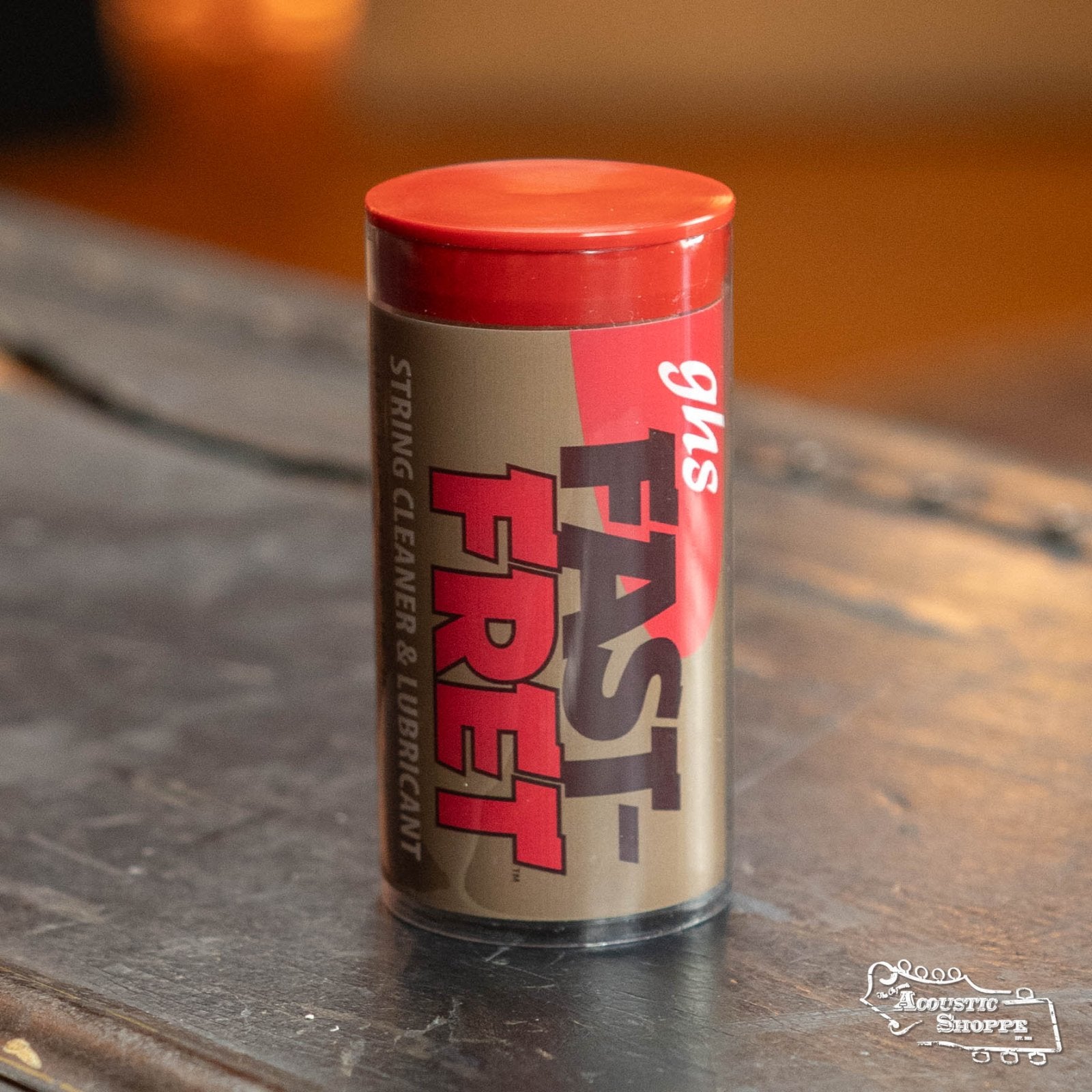 A container of GHS Strings A87 Fast Fret String Cleaner/Lubricant with a red cap rests on a dark wooden surface, its branding visible and emphasizing its ability to prolong string life.