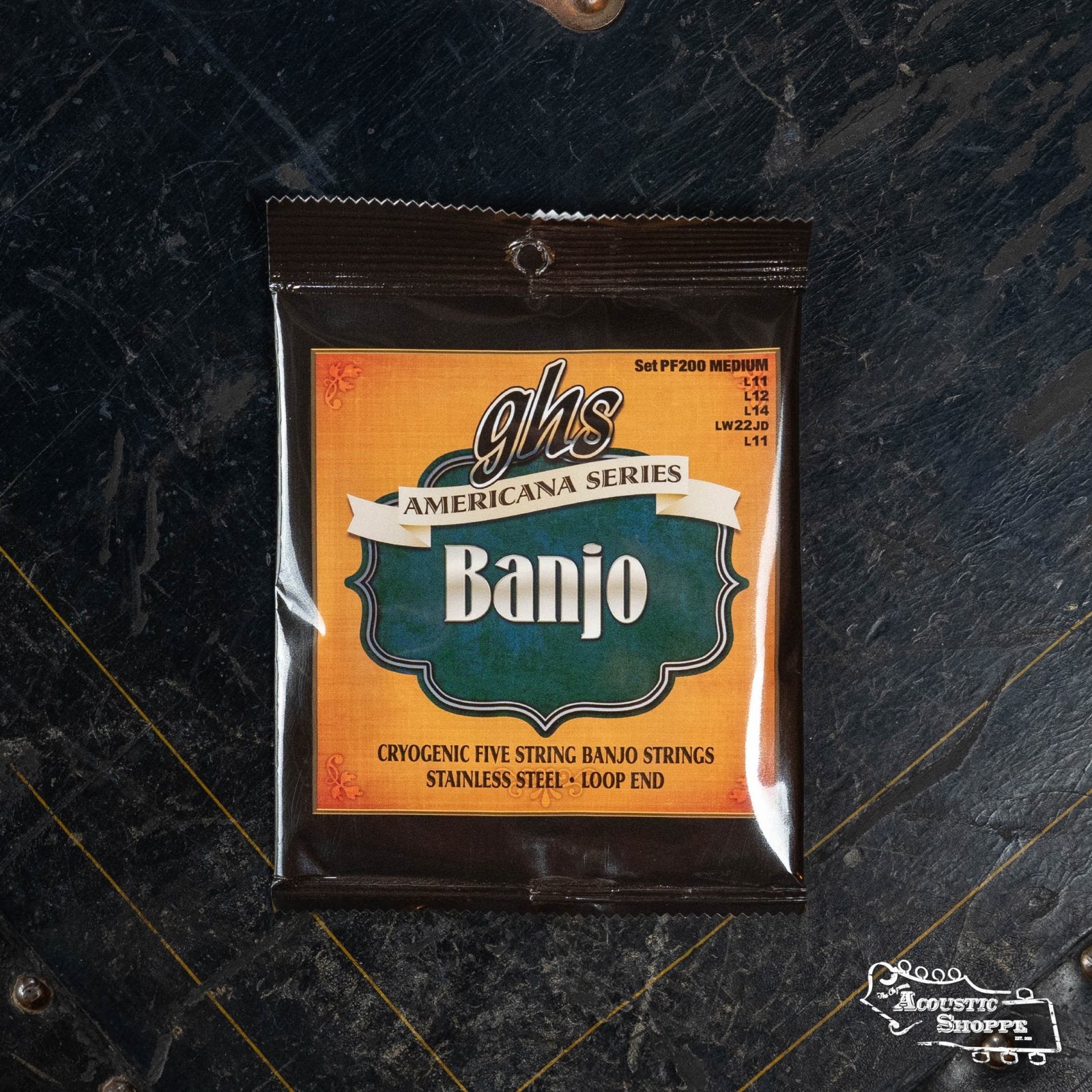 A packet of GHS Strings PF200 Stainless Steel 5-String Banjo - Medium rests on a dark, textured surface. The vibrant orange and green packaging displays product details and the GHS Strings logo.