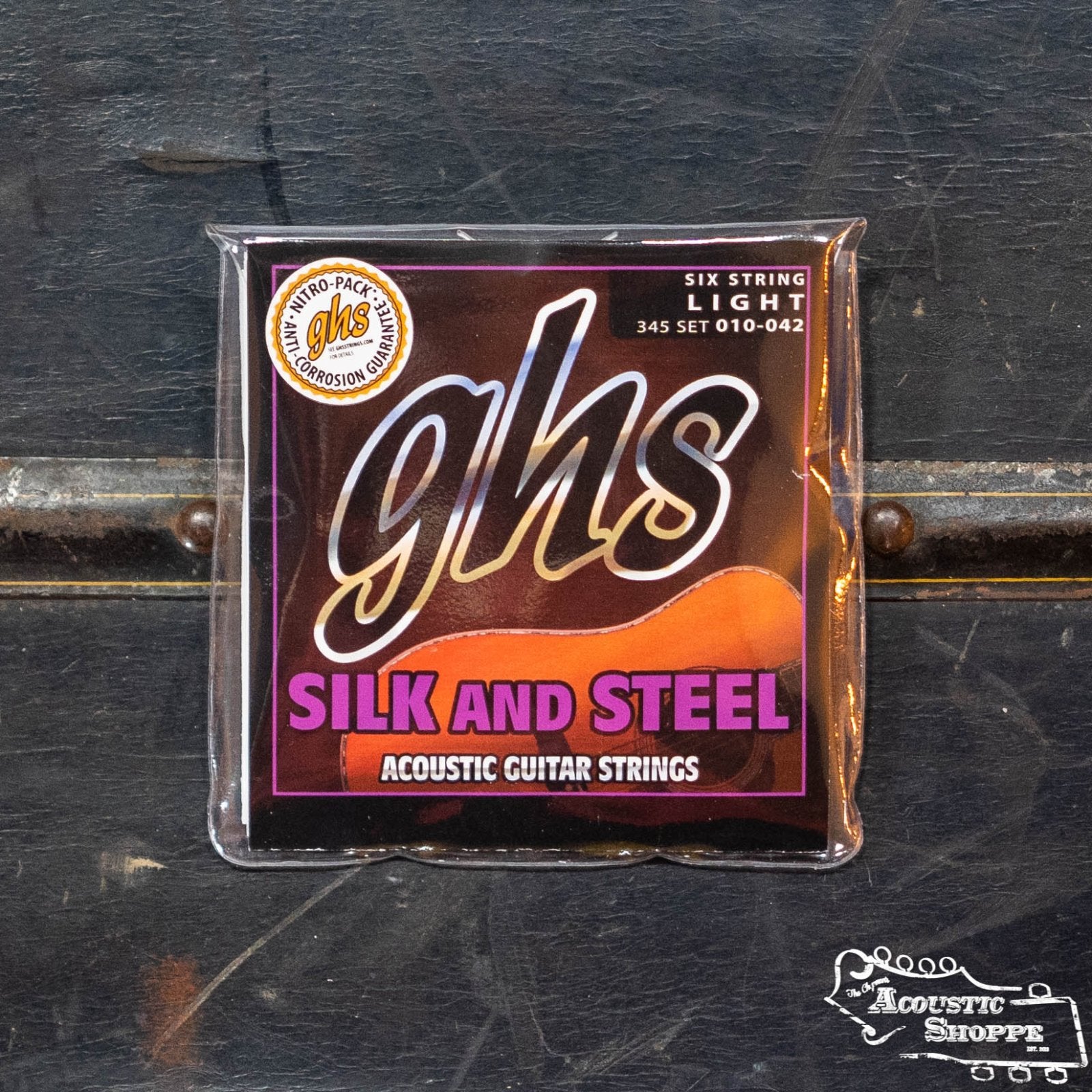 A package of GHS 345 Silk and Steel Acoustic Guitar Strings - Light by GHS Strings rests on a dark, textured surface, with The Acoustic Shoppe logo in the bottom right, promising a warm, mellow tone.