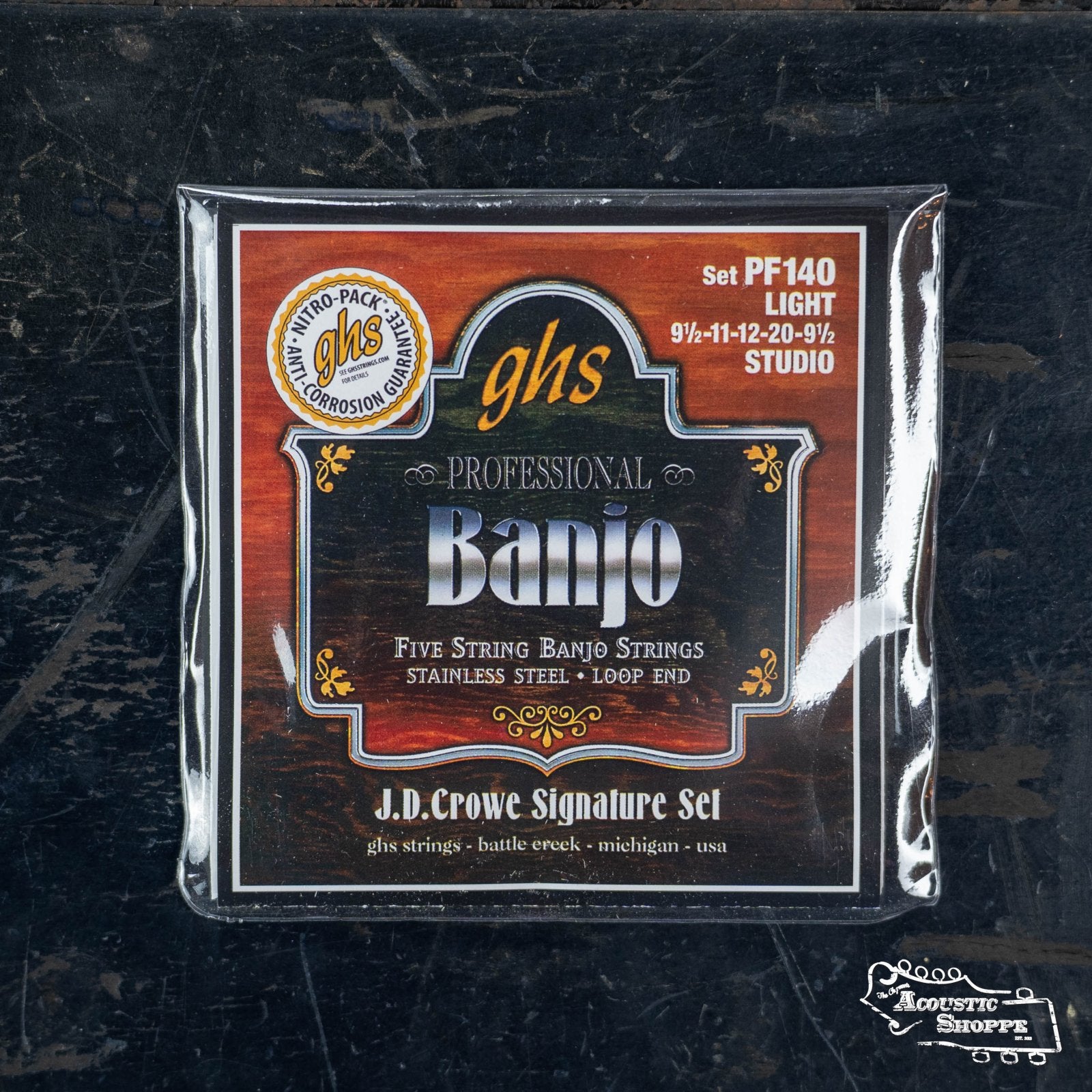 A packet of GHS Strings J.D. Crowe Signature Studio 5-string Banjo Stainless Steel Strings (PF140), featuring light gauges and Lock Twist loop ends, sits on a dark, worn surface.