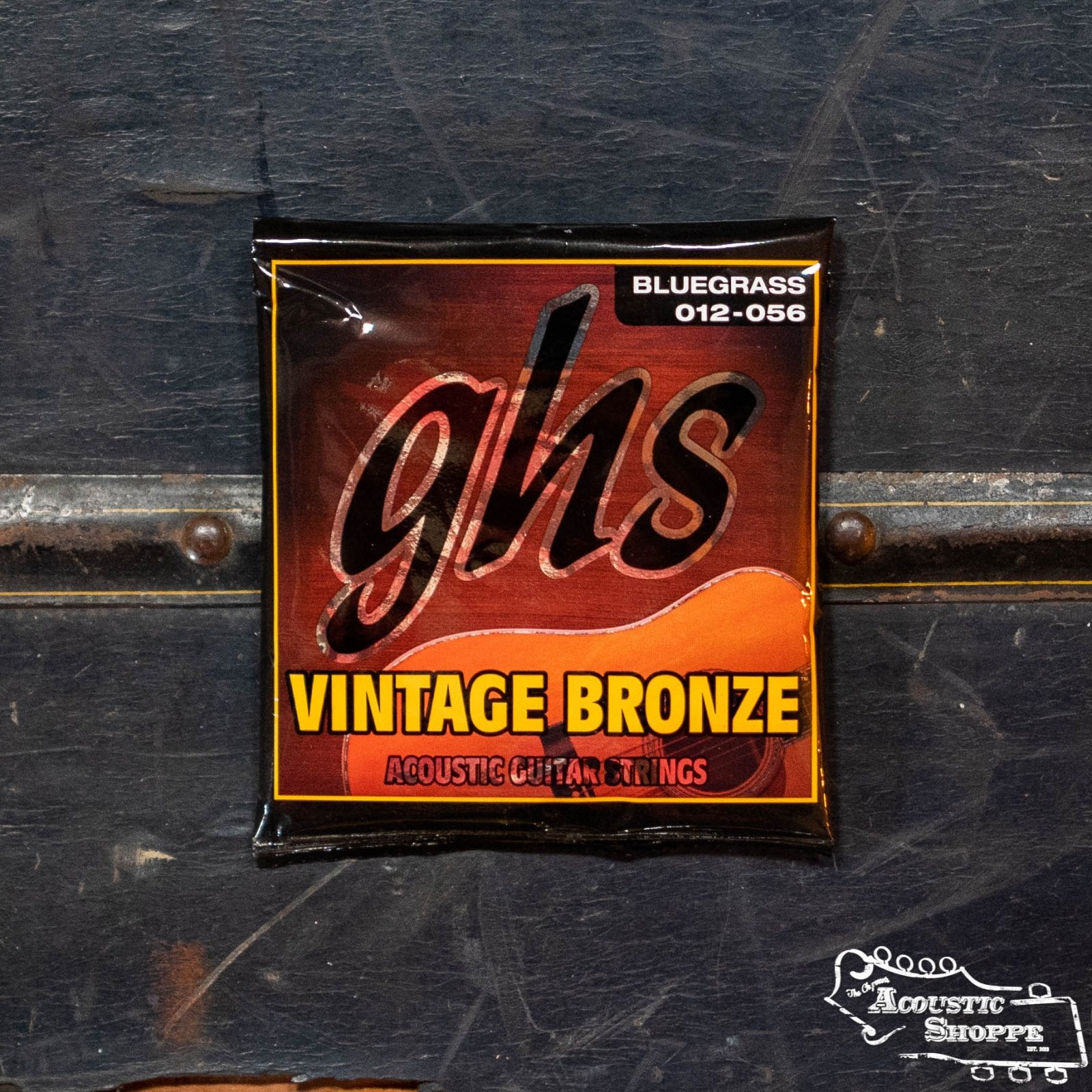 A pack of GHS Strings Vintage Bronze Bluegrass Acoustic Guitar Strings (VN-B) sits on a dark, textured surface, with The Acoustic Shoppe logo visible in the lower right corner.
