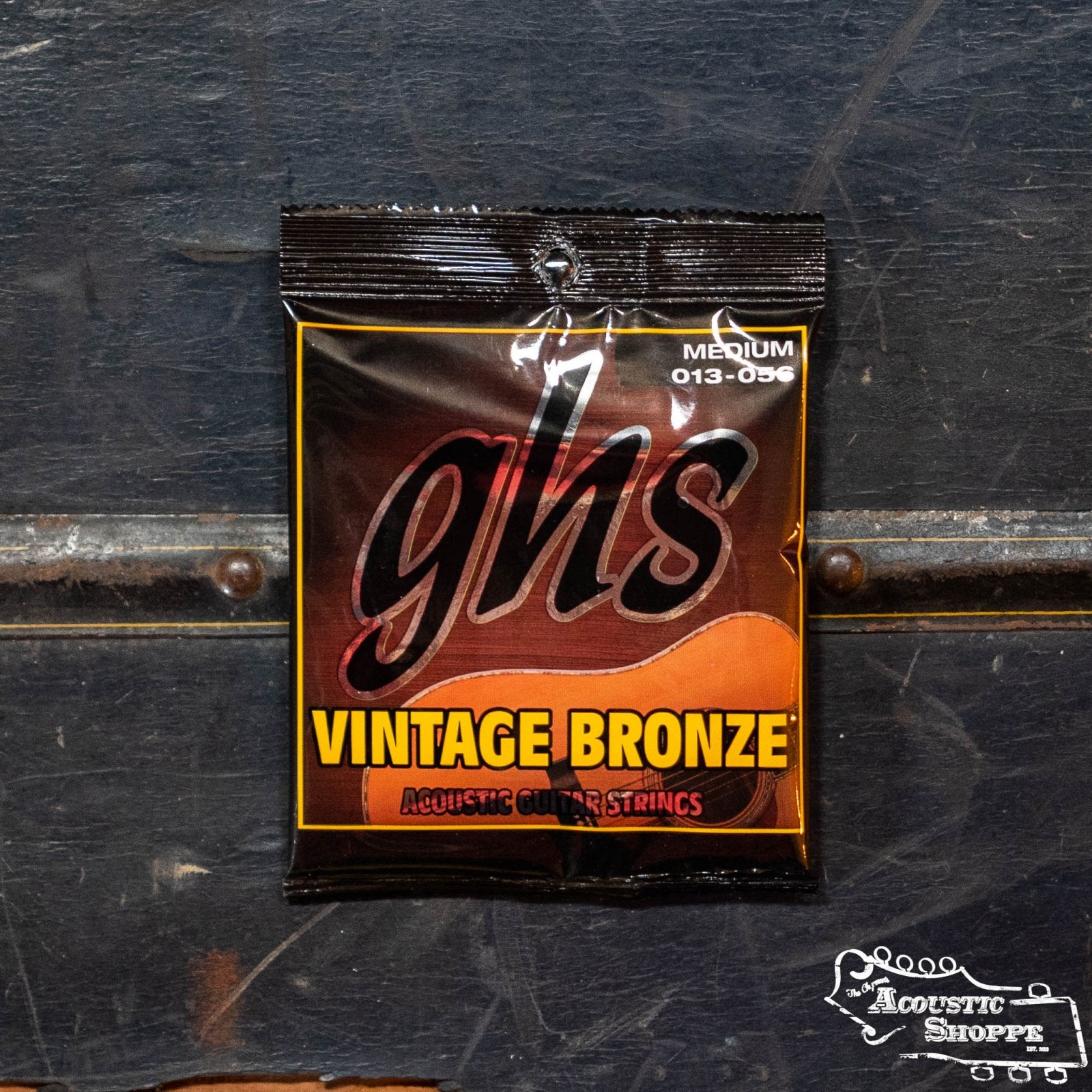 A pack of GHS Strings Vintage Bronze Medium Acoustic Guitar Strings (VN-M) rests on a dark textured surface, with the black packaging featuring bronze and yellow text plus a small The Acoustic Shoppe logo in the lower right corner.