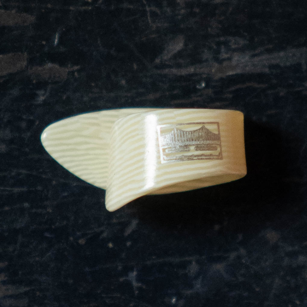 A large, cream-colored Golden Gate GP-10 Ivoroid Thumb Pick, featuring a textured surface and a printed bridge image on the front, rests on a dark, worn surface.