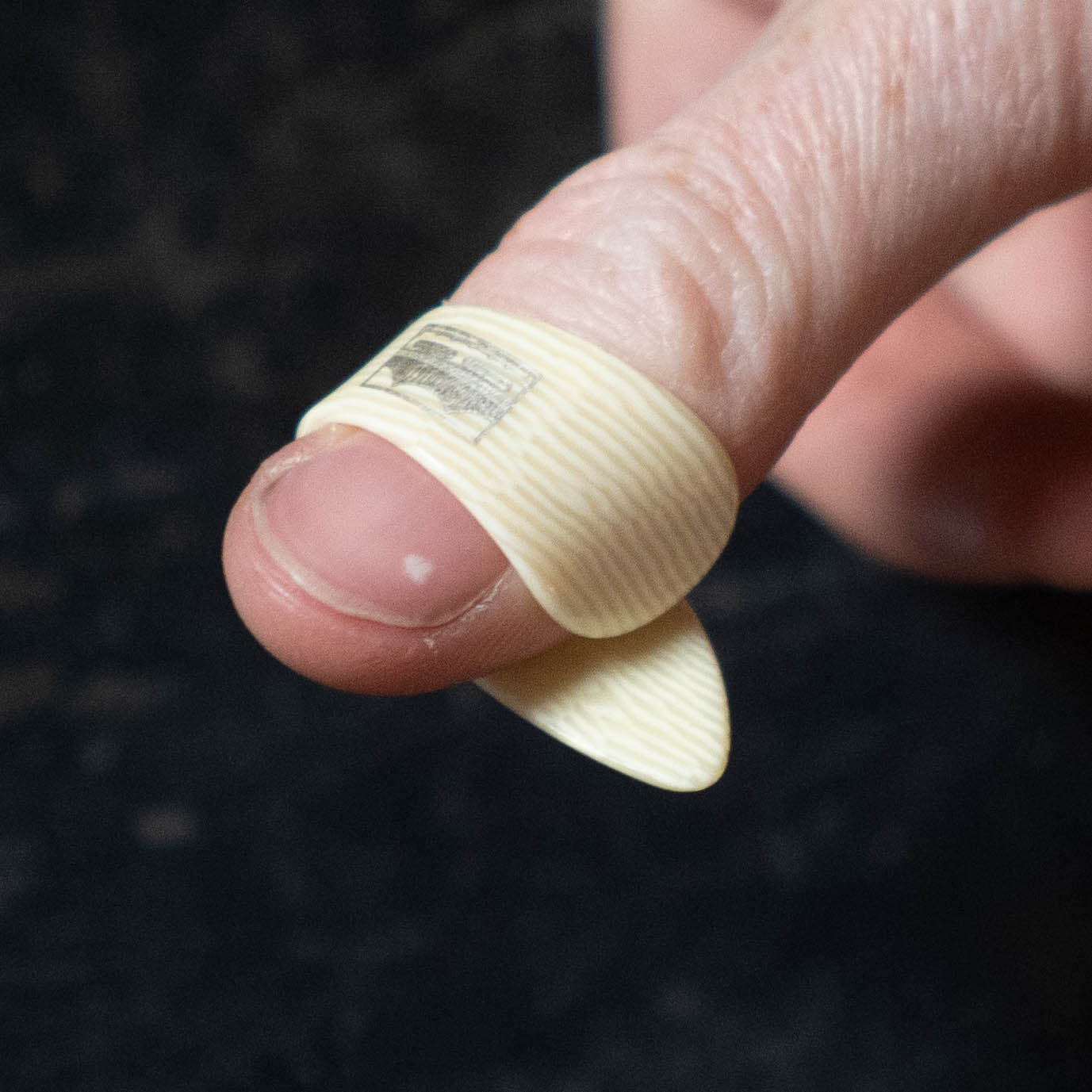 A close-up of a person's finger wearing a Golden Gate GP-10 Ivoroid Thumb Pick - LARGE by Golden Gate, cream-colored, against a dark background.