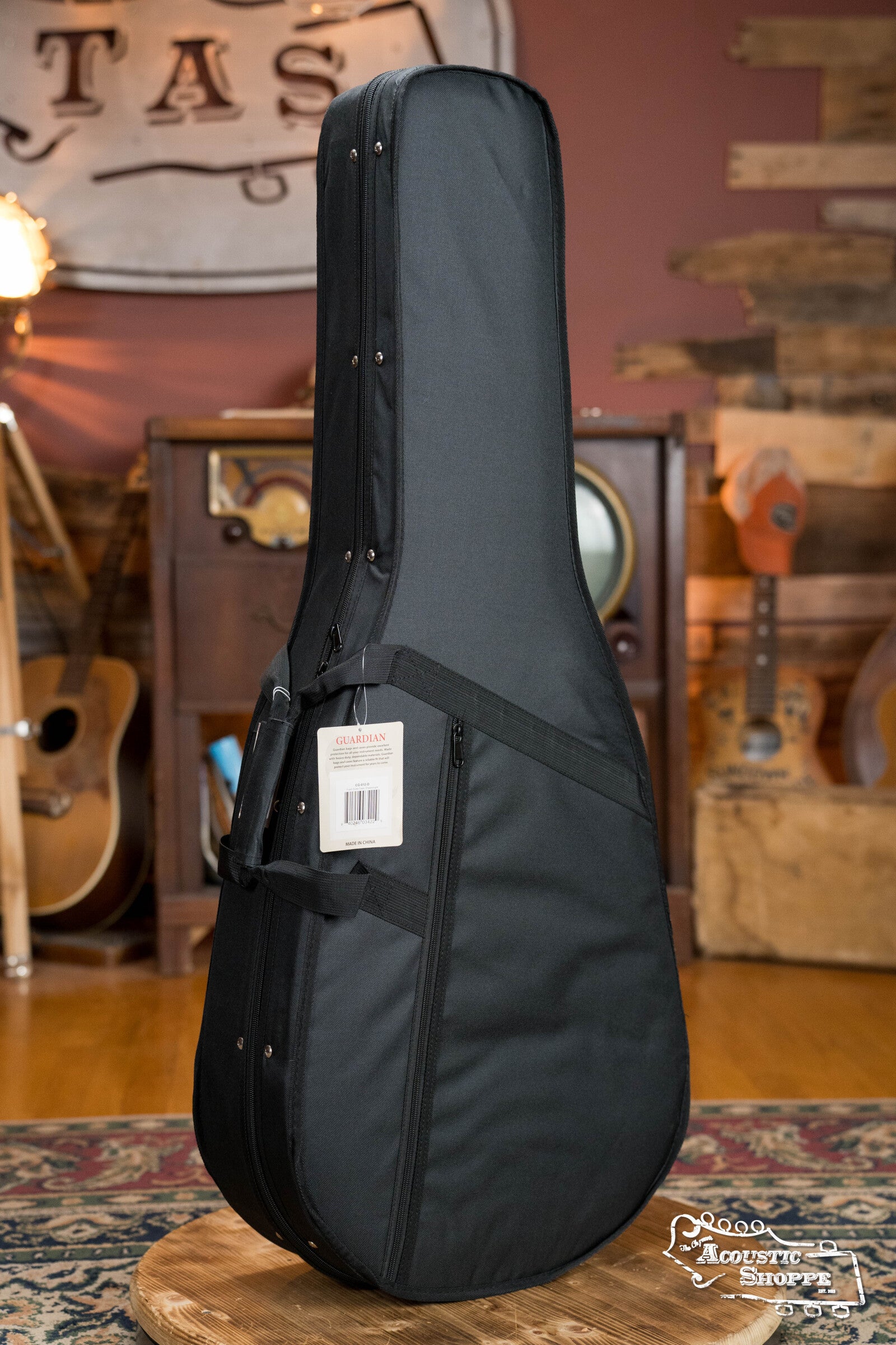 The Guardian CG-012-D Foam Guitar Case Dreadnought by Guardian, featuring high-density foam padding and a white label with red text on the handle, stands on a wooden surface in a guitar-filled, vintage music room. Perfect fit for dreadnought guitars.