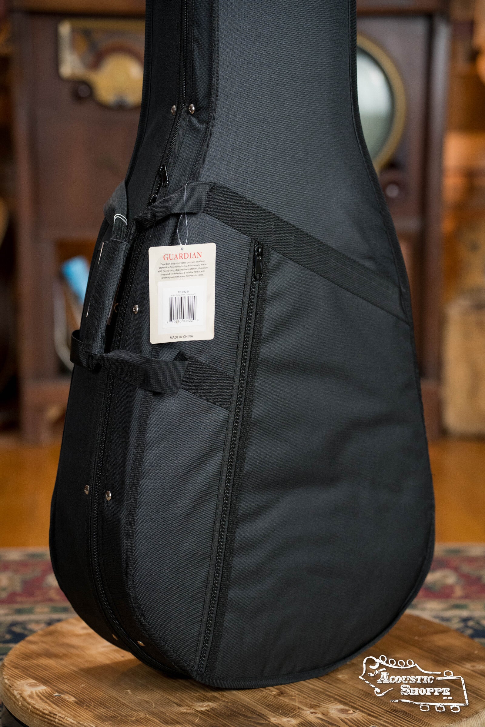 The Guardian CG-012-D Foam Guitar Case Dreadnought is shown upright on a round wooden stool. It features high-density foam padding and a white Guardian tag, with The Acoustic Shoppe logo in the bottom right corner.