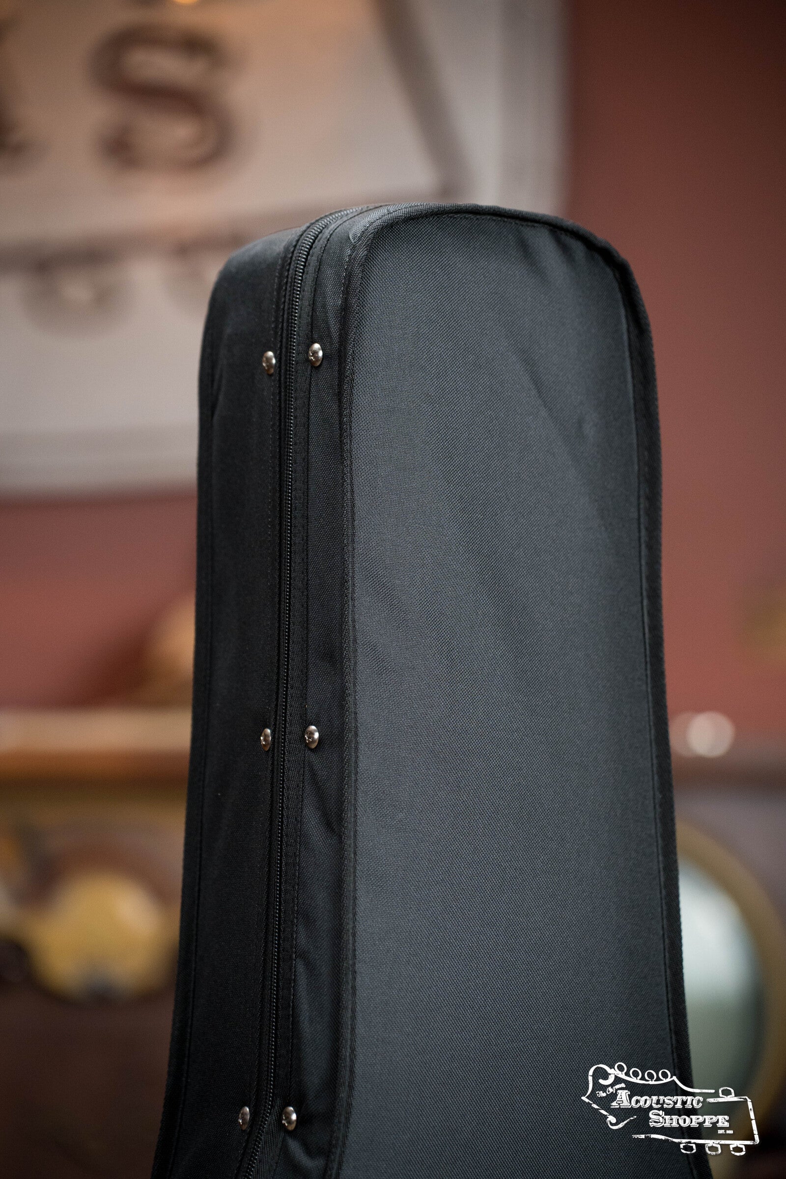 A close-up of the Guardian CG-012-D Foam Guitar Case Dreadnought by Guardian stands upright, showcasing its black padded design with silver rivets. The background is blurred, and The Acoustic Shoppe logo appears in the bottom right corner.
