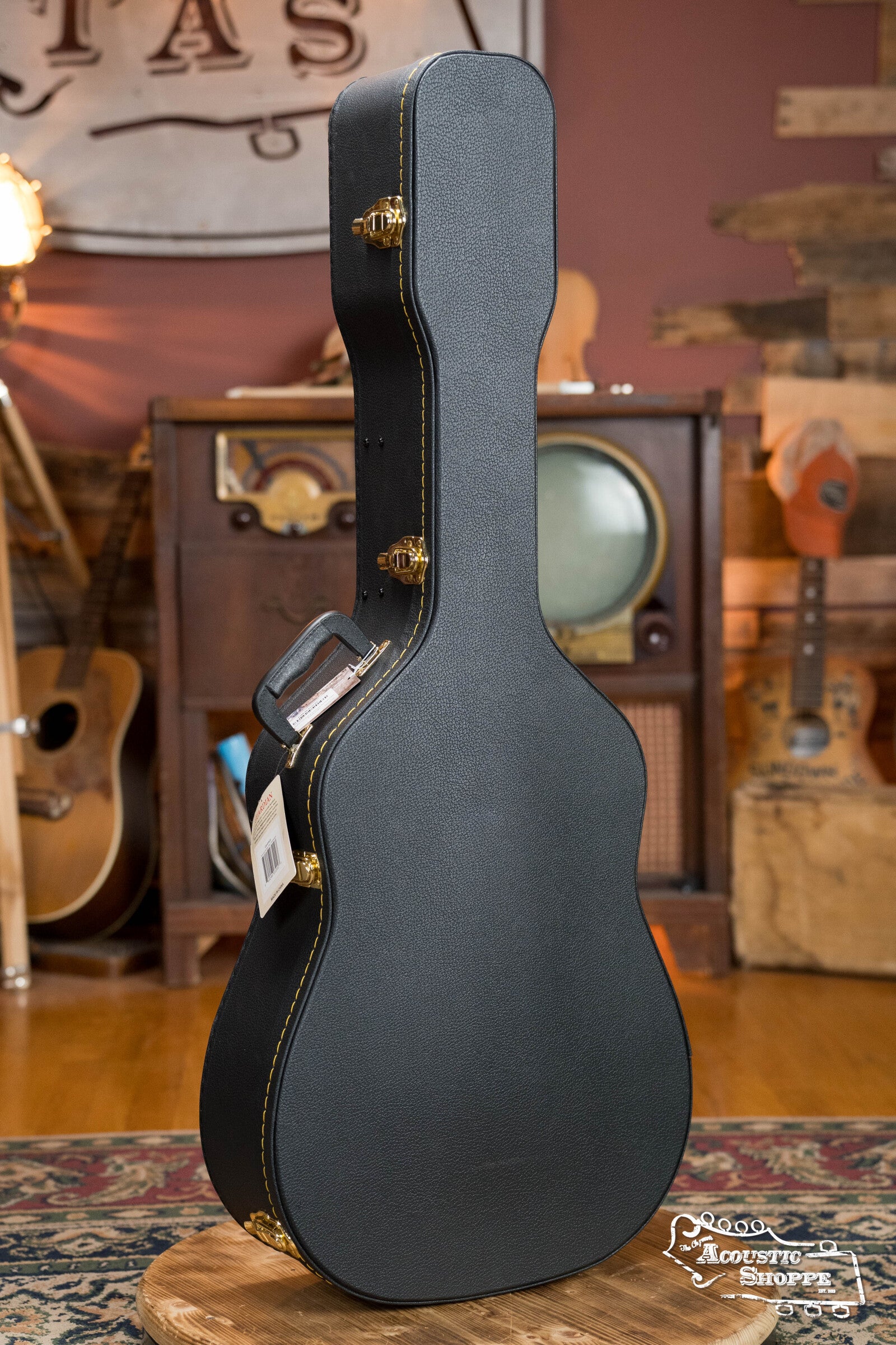A Guardian CG-018-D Economy Archtop Case Dreadnought by Guardian, in black with gold latches, stands upright on a round rug in a cozy, music-themed room featuring wooden furniture, guitars, and a vintage TV.