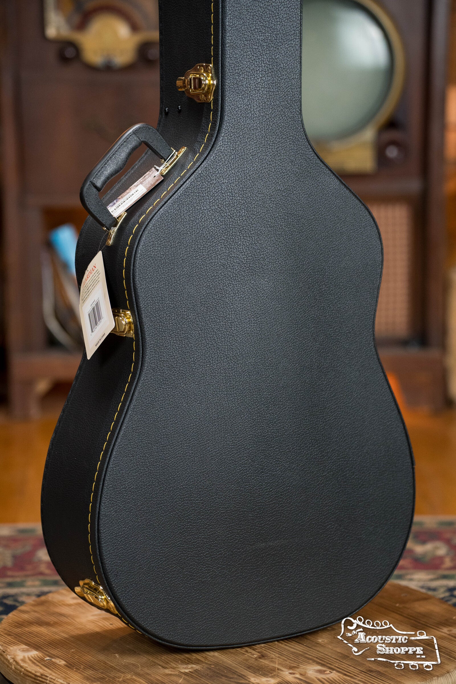 The Guardian CG-018-D Economy Archtop Case Dreadnought by Guardian stands upright on a wooden surface, featuring black finish, gold hardware, yellow stitching, a handle tag, and the Acoustic Shoppe logo in white at the bottom right corner.