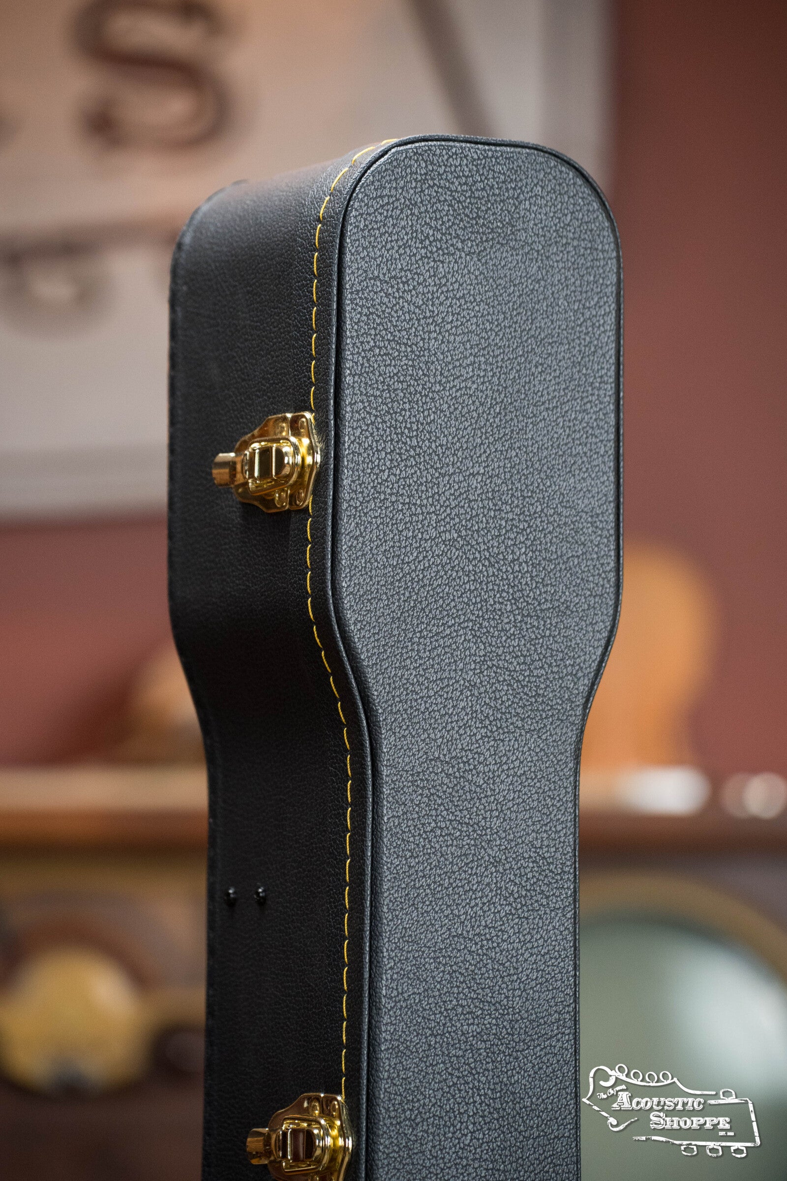 Close-up of a Guardian CG-018-D Economy Archtop Case for Dreadnought guitars in black with yellow stitching and gold latches, standing upright. A blurred background reveals part of a sign and indistinct objects.