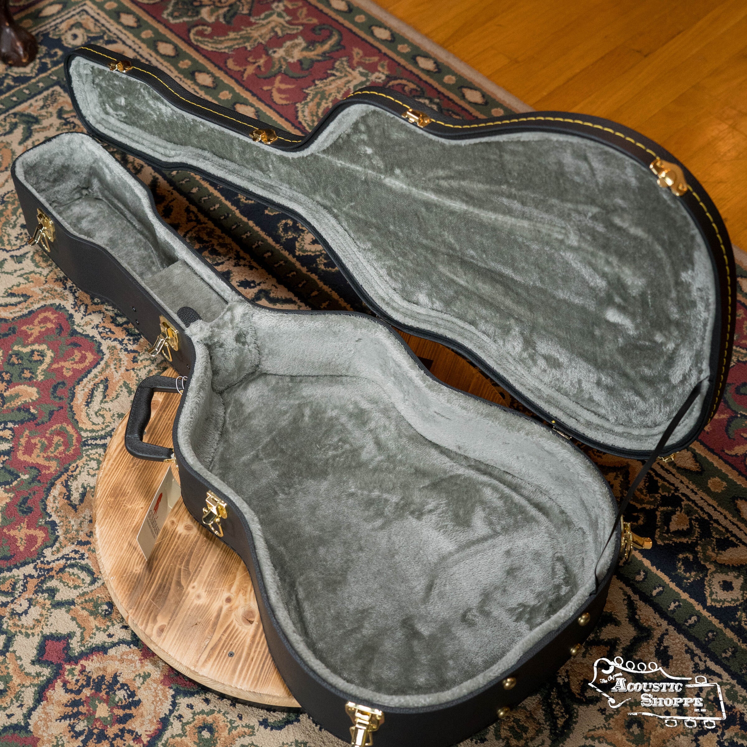 A Guardian CG-018-D Economy Archtop Dreadnought case by Guardian, featuring a black exterior, gold hardware, gray plush interior, sits open and empty on a round wooden table over a patterned rug.
