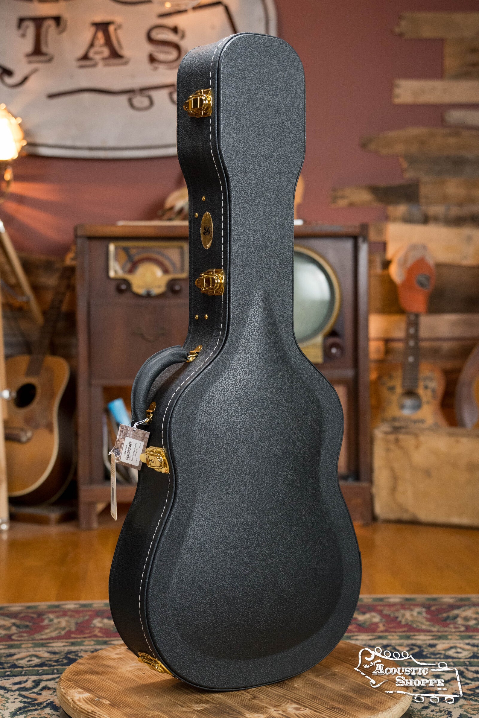 A Guardian CG-044-OOO Vintage OOO Hardshell Case by The Music Link with gold latches stands upright on a round wooden table in a cozy room adorned with vintage OOO guitars, an old TV, and rustic wooden accents.