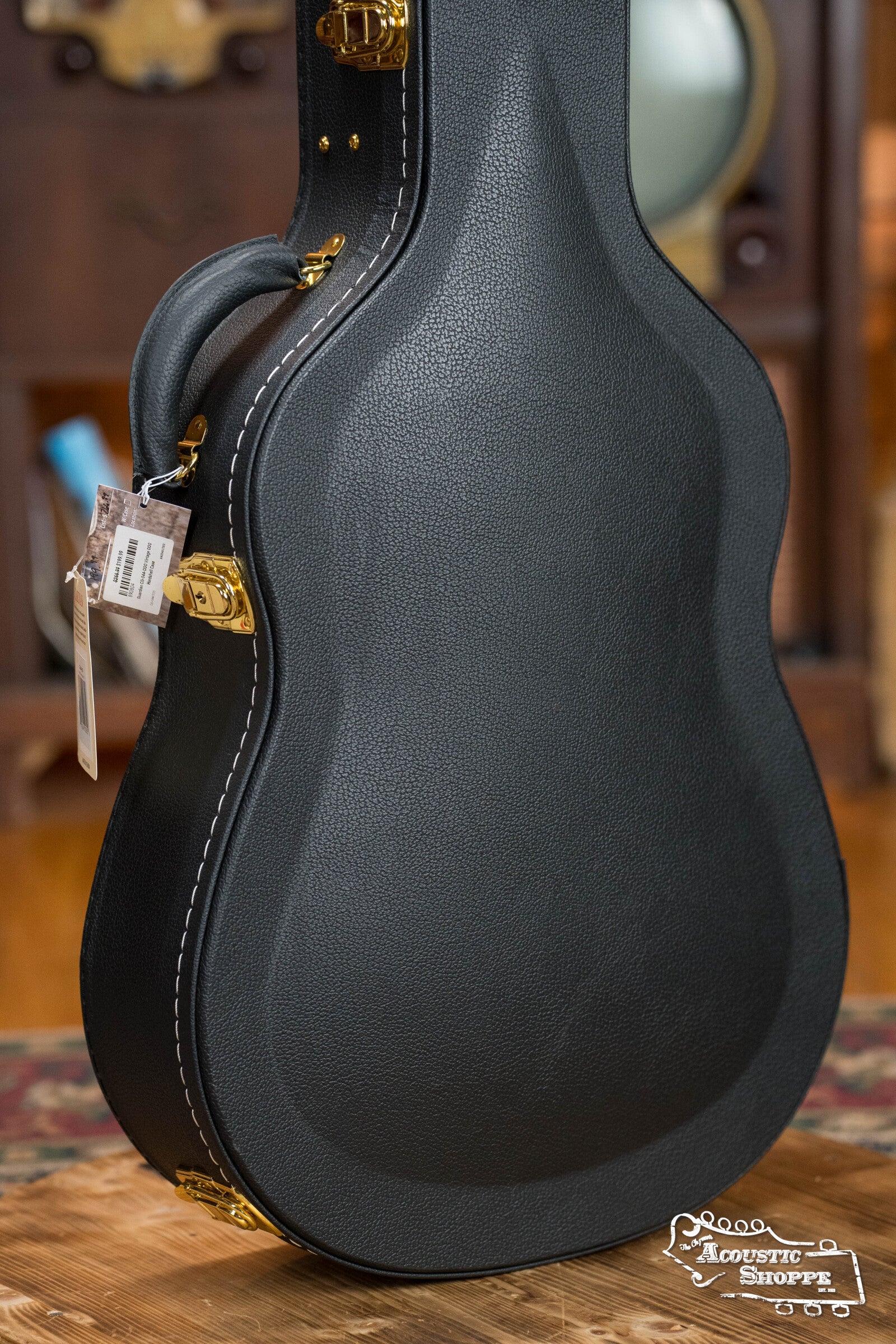 A black, textured Guardian CG-044-OOO Vintage OOO Hardshell Case by The Music Link is upright on a wooden surface with gold latches, white stitching, a tag on the handle, and an Acoustic Shoppe logo in the bottom right corner.