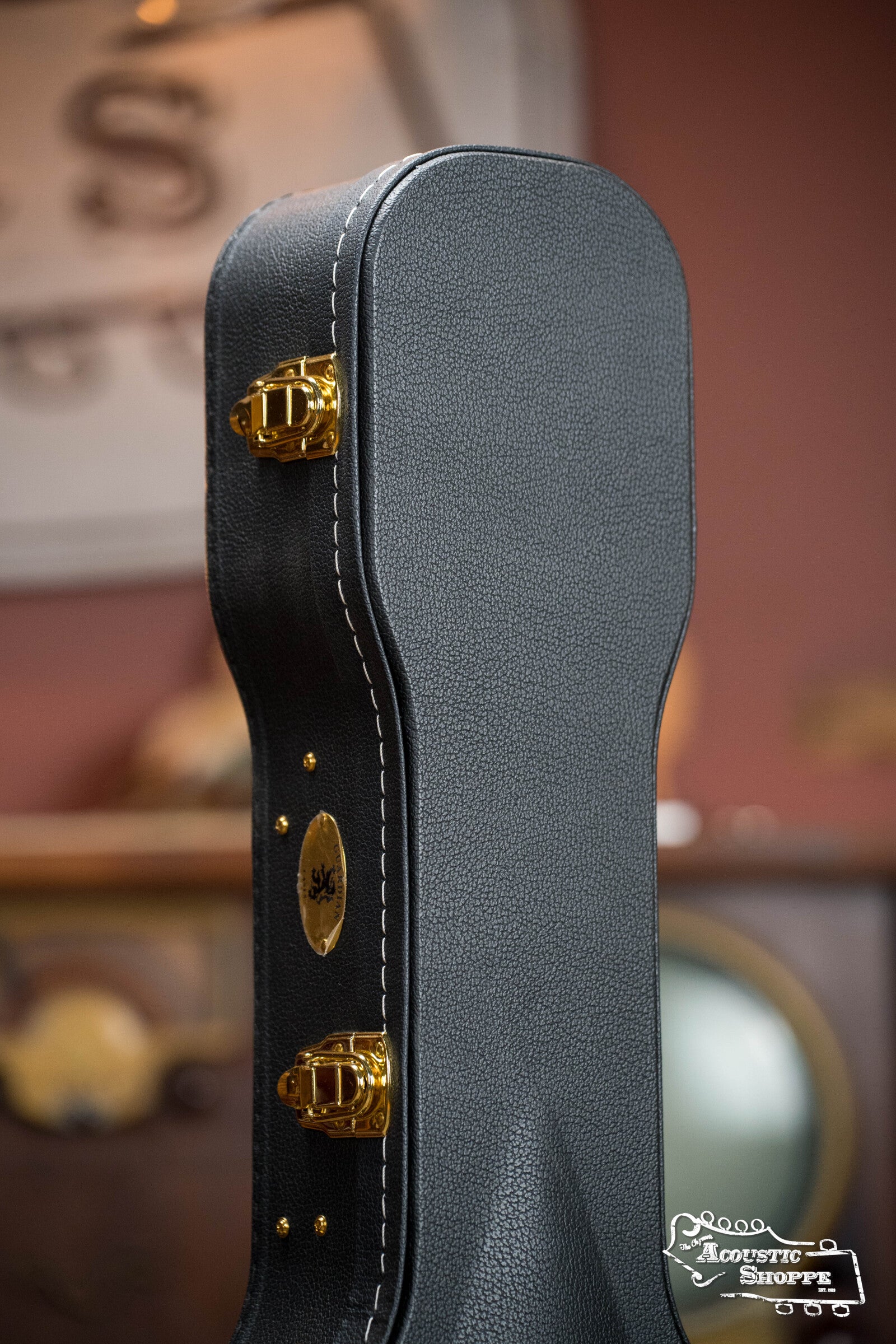 A close-up of The Music Link Guardian CG-044-OOO Vintage OOO Hardshell Case in textured black with gold hardware and white stitching, upright. A blurred background with a round mirror and vintage decor. Acoustic Shoppe logo appears in the corner.