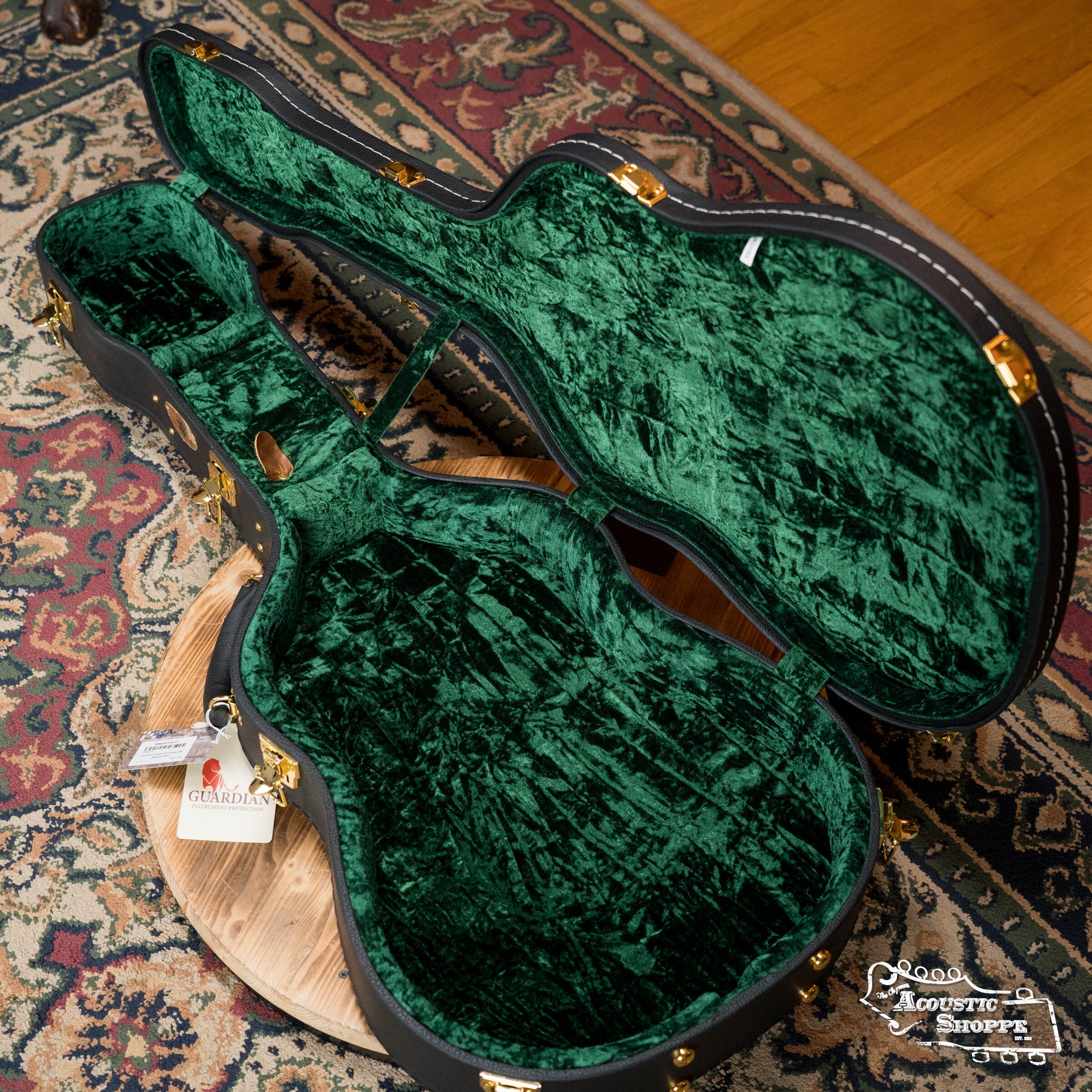 An open, empty The Music Link Guardian CG-044-OOO Vintage OOO Hardshell Case with plush green interior sits on a round wooden stool over a patterned rug, with a Guardian tag attached to the handle.