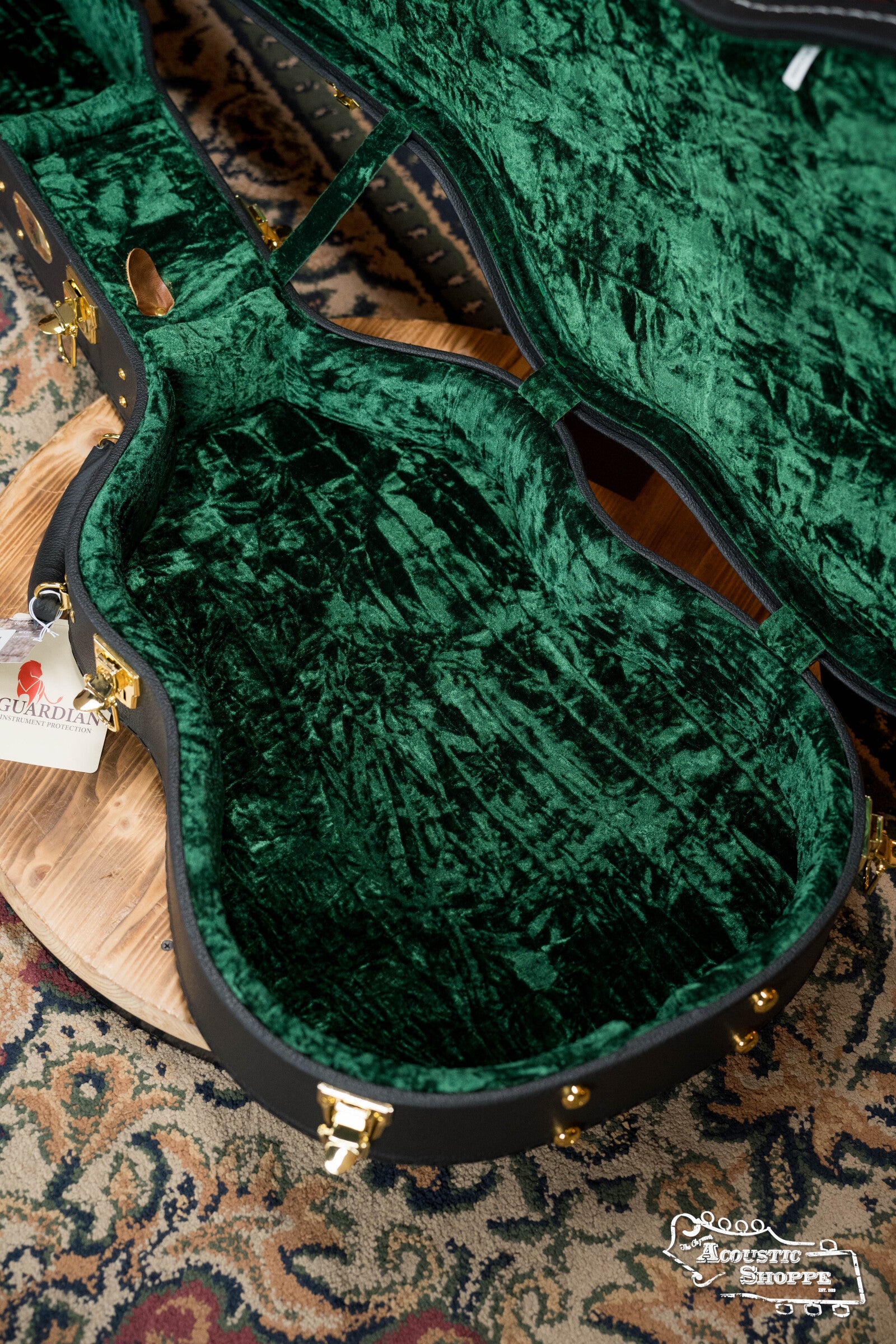A Guardian CG-044-OOO Vintage OOO Hardshell Case by The Music Link, with plush emerald-green interior, gold latches, and a woodgrain Guardian tag, sits open on a patterned rug. Part of a circular wooden stand is visible underneath.