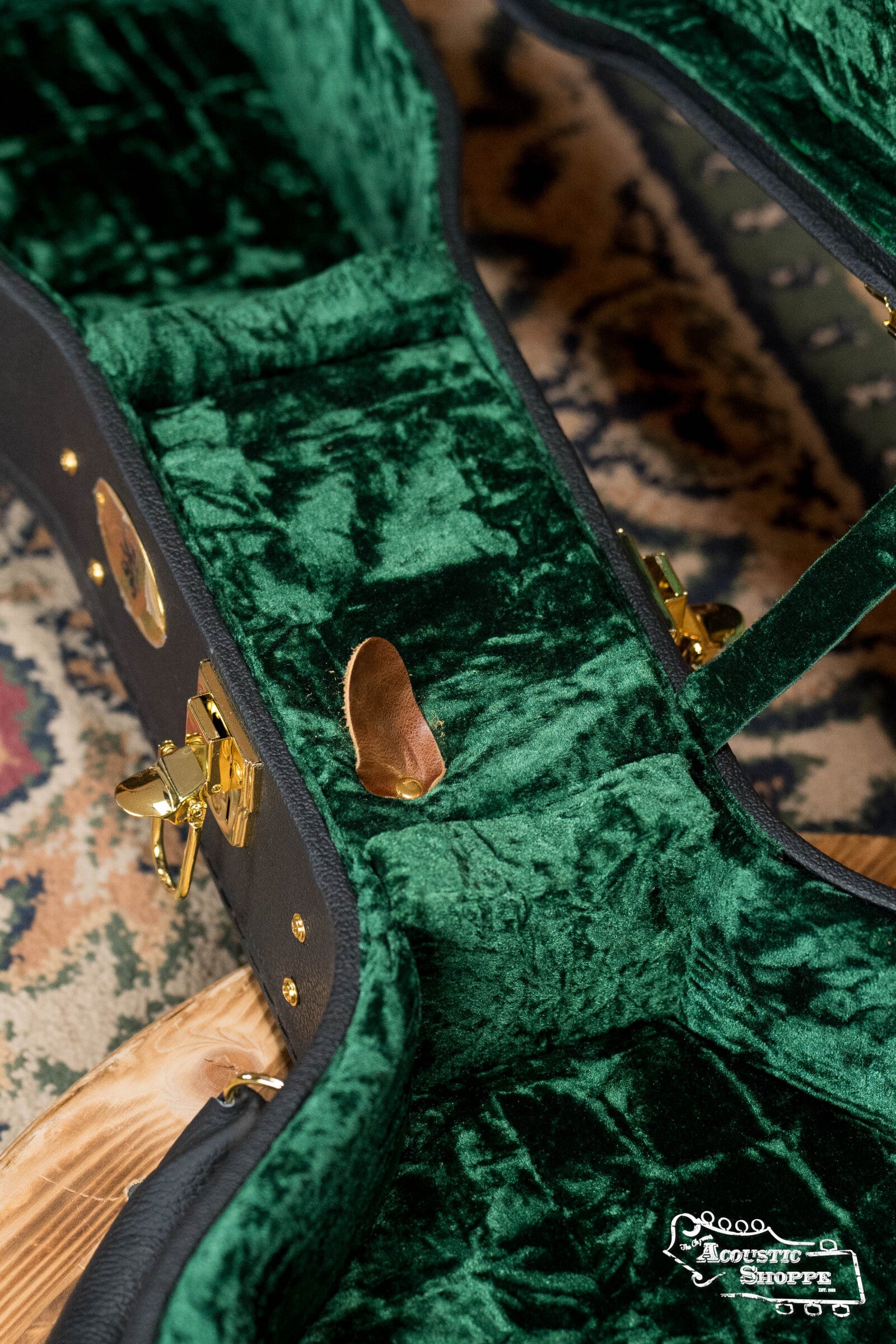 A brown guitar pick sits in a plush, green velvet-lined Guardian CG-044-OOO Vintage OOO Hardshell Case by The Music Link with gold hardware, atop a patterned rug. The Acoustic Shoppe logo appears in the corner.