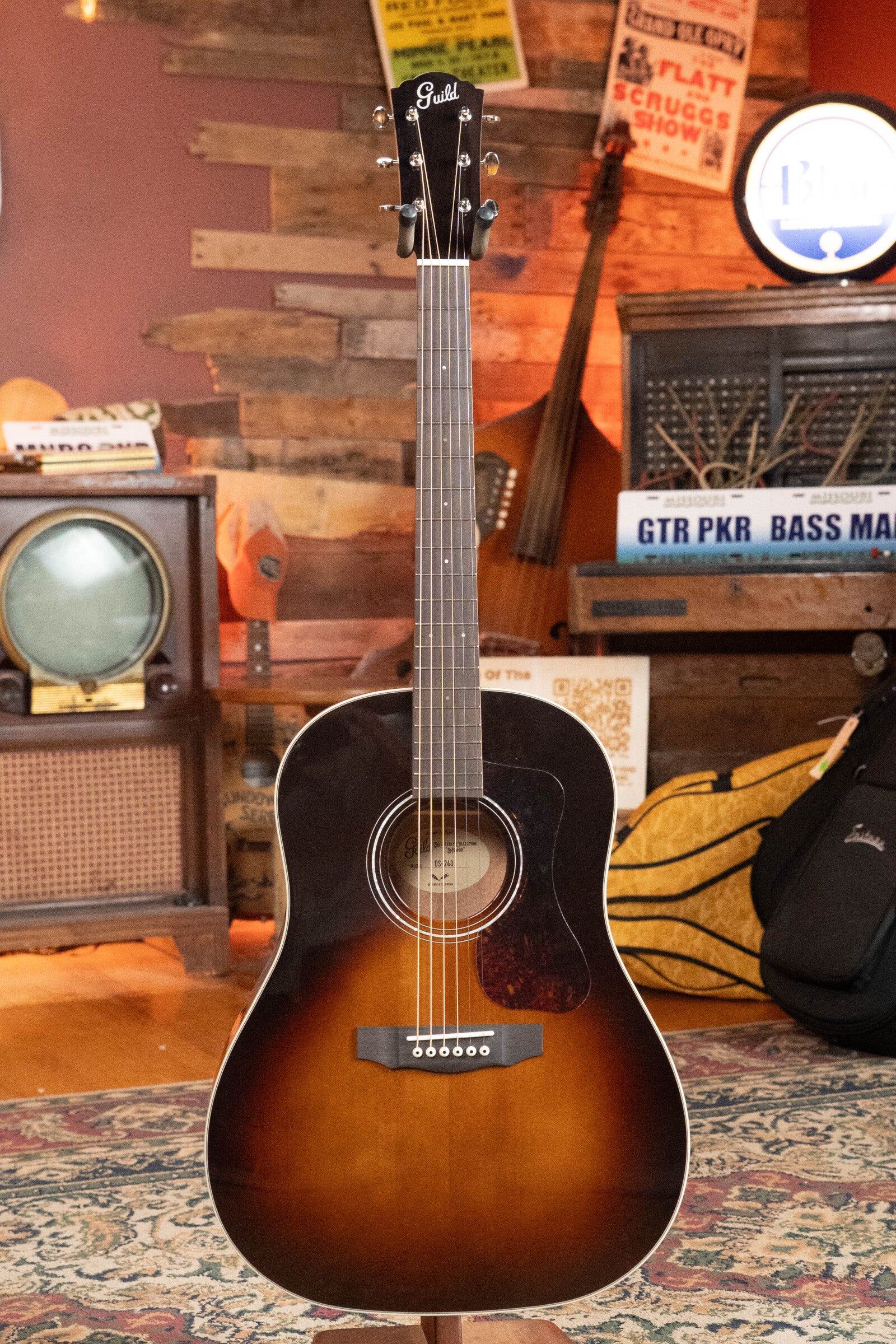 A Guild DS-240 Memoir Sitka/Mahogany Vintage Sunburst Slope Shoulder Dreadnought Acoustic Guitar by Guild Guitars stands on a patterned rug in a cozy vintage room filled with old signs, a retro TV, a woven basket, and music gear.