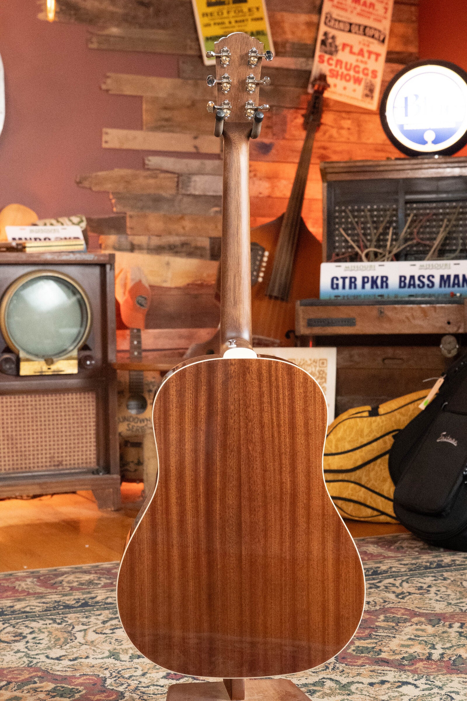 The Guild DS-240 Memoir Sitka/Mahogany Vintage Sunburst Slope Shoulder Dreadnought Acoustic Guitar by Guild Guitars stands upright with its back to the camera in a cozy, vintage-inspired room filled with retro signs, a TV, radio, and musical gear.