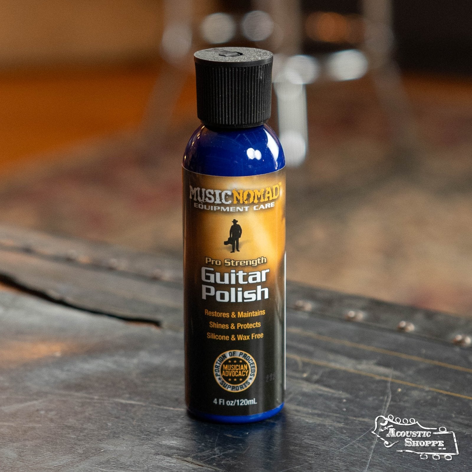 A blue bottle of Music Nomad Pro-Strength Formula Guitar Polish (MN101) sits on a wooden surface, its label emphasizing restoration of guitar shine. The Acoustic Shoppe logo is visible in the corner.