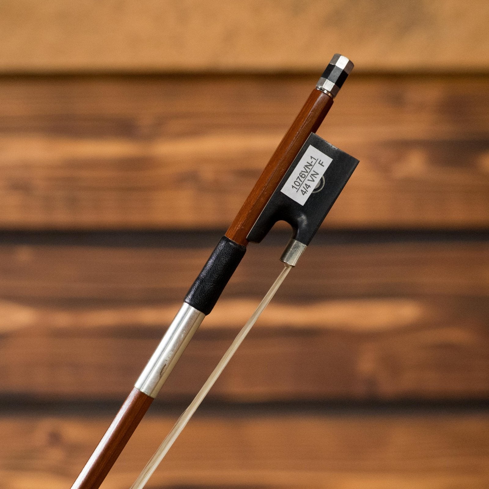The Howard Core 1076VN 4/4 Violin Bow features a Brazilwood octagonal stick, ebony frog, and silver fittings, highlighted in a close-up view against a softly blurred wooden background.