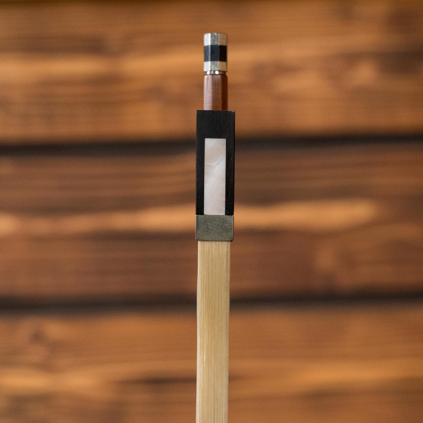 Close-up of the Howard Core 1076VN 4/4 Violin Bow’s ebony frog and octagonal Brazilwood stick, featuring a rectangular mother-of-pearl inlay, with a blurred wooden background.