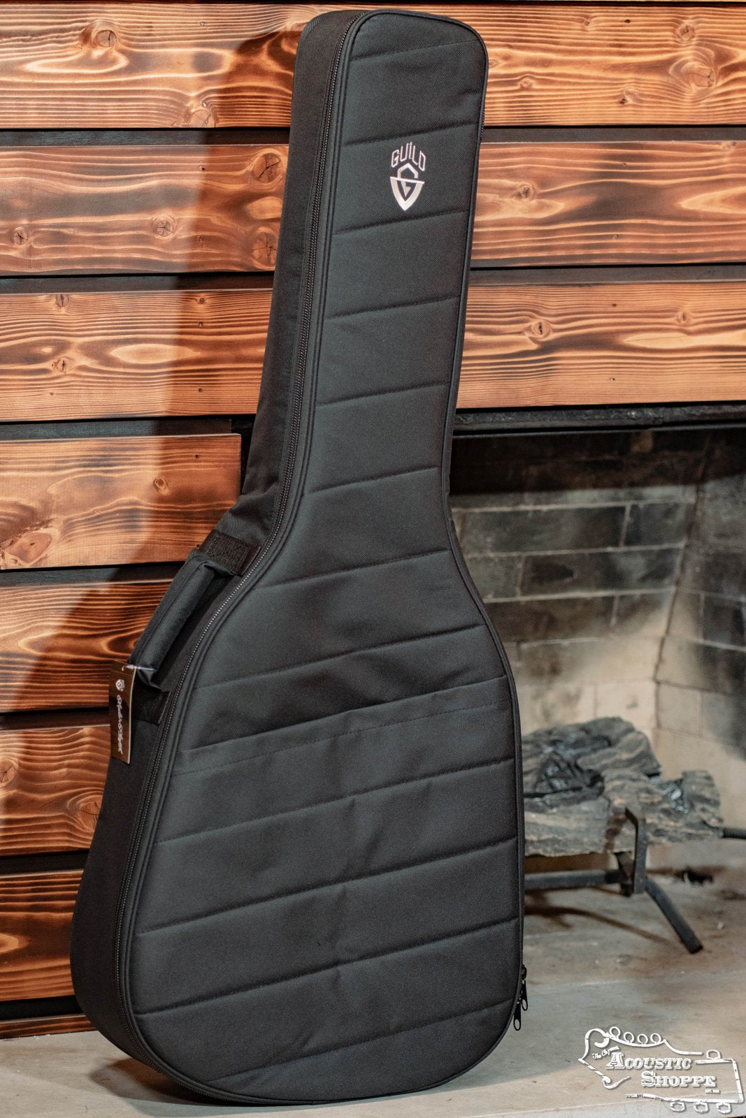 The Guild Deluxe Acoustic Gig Bag - Jumbo by Guild Guitars, shown upright by a rustic wood wall and stone fireplace, is perfect for protecting your jumbo-size acoustic guitars and other instruments.