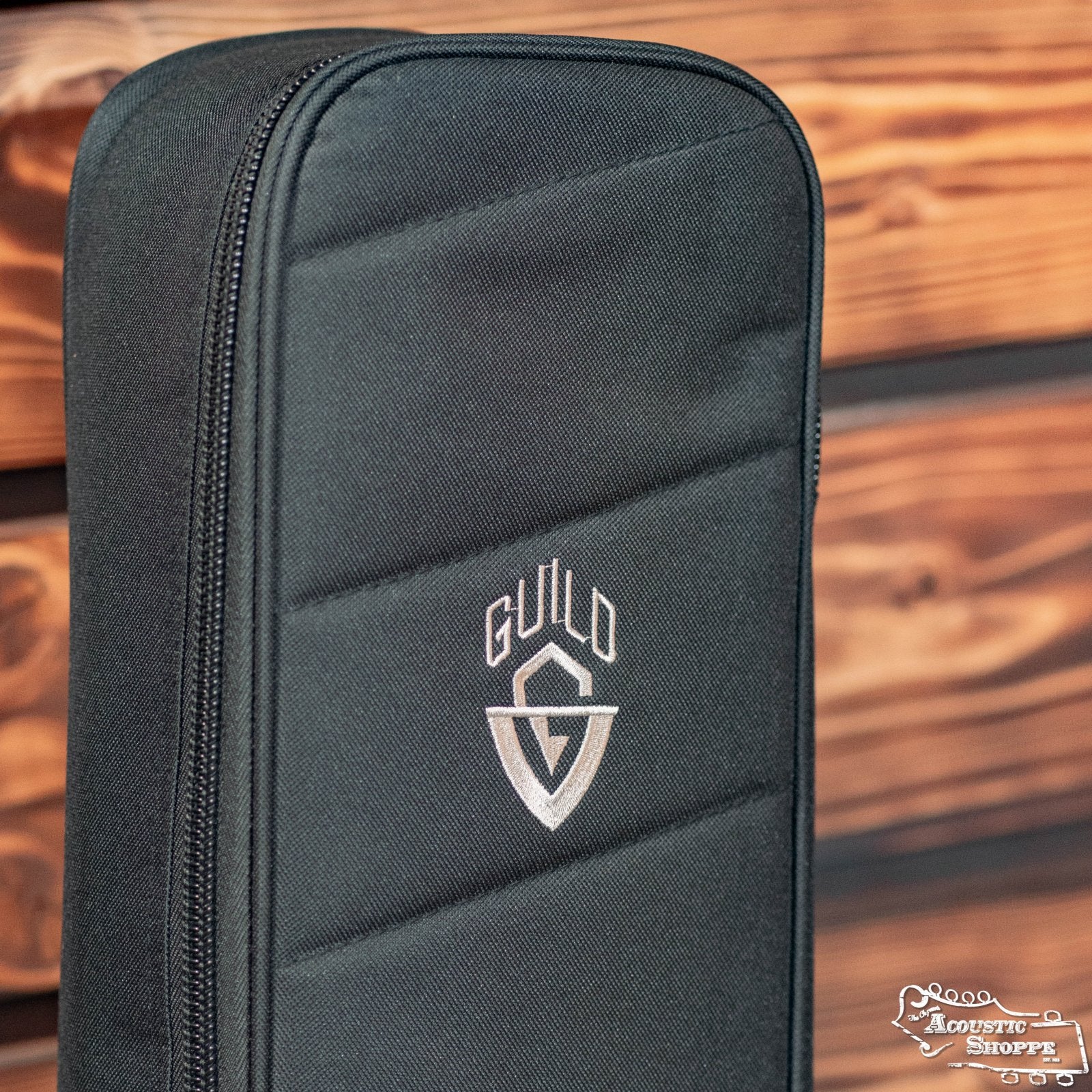 A close-up of the Guild Deluxe Acoustic Gig Bag - Jumbo by Guild Guitars, featuring a white embroidered logo and visible stitching and zippers, set against a wooden background.
