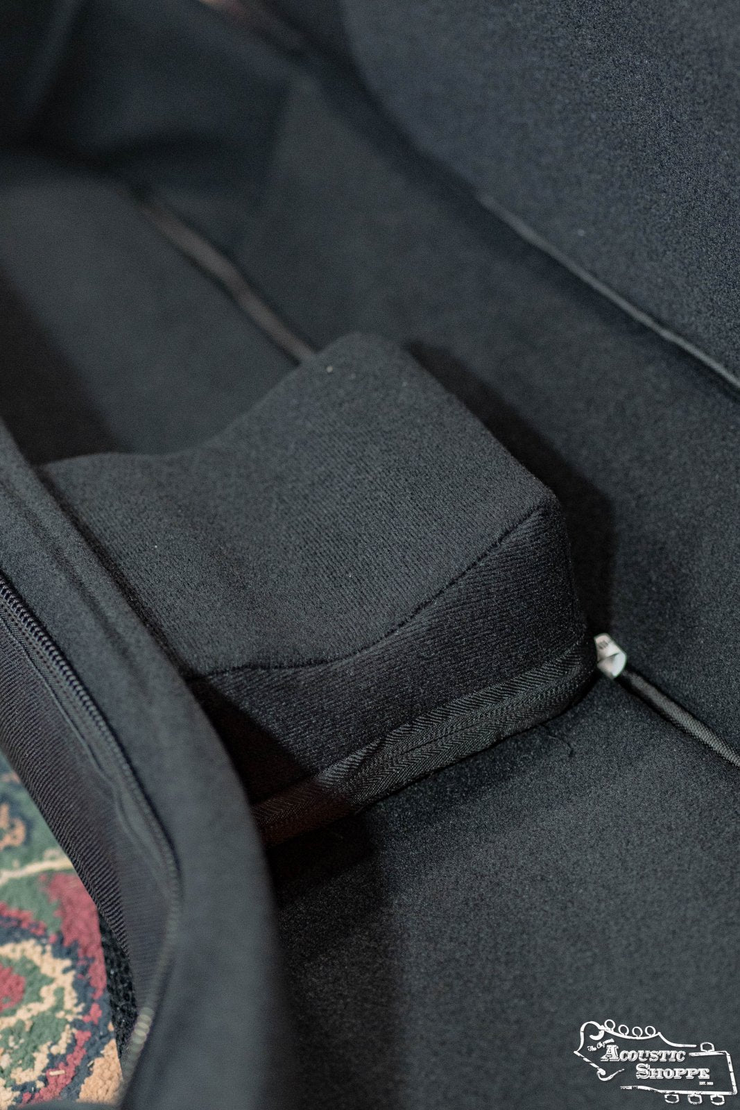 A close-up of the Guild Deluxe Acoustic Gig Bag - Jumbo by Guild Guitars shows its soft lining and raised support block for premium protection. A patterned rug and Guild Guitars logo are partially visible in the background.