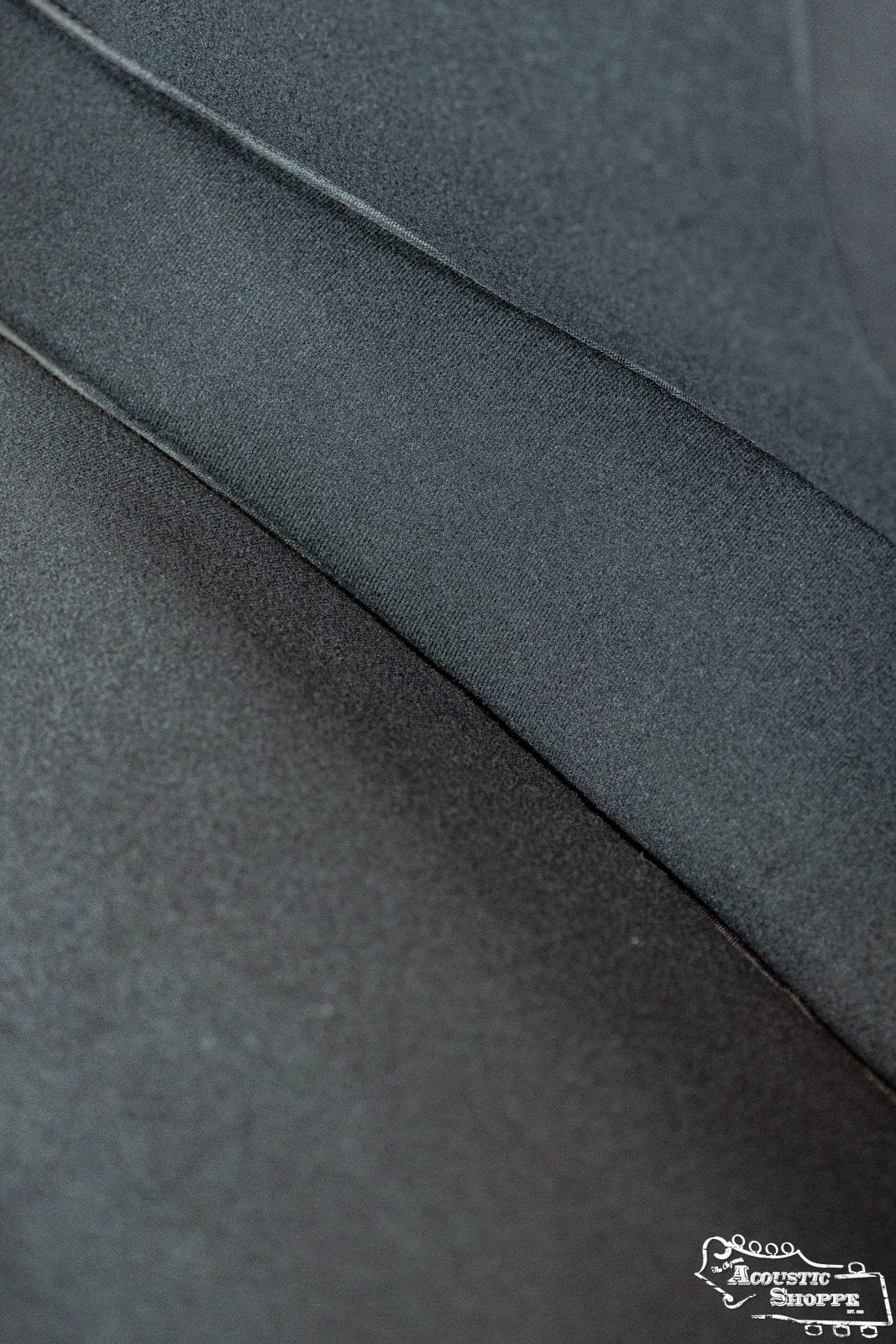 Close-up of black fabric with a diagonal seam and visible texture, reflecting the quality of Guild Guitars. The soft-focus image features the Guild Deluxe Acoustic Gig Bag - Jumbo with a white logo in the bottom right corner.