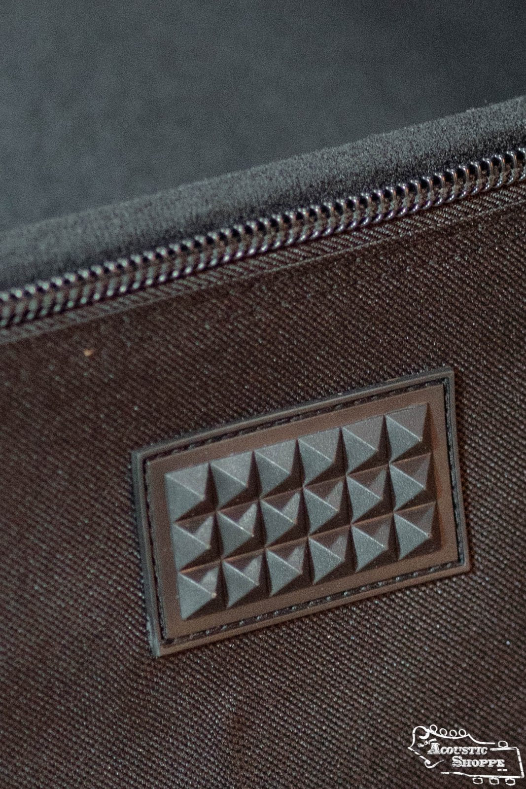 Close-up of the Guild Deluxe Acoustic Gig Bag - Jumbo by Guild Guitars, featuring brown fabric, a visible black zipper, and a rectangular patch with raised pyramid studs. The Acoustic Store logo is in the bottom right corner.