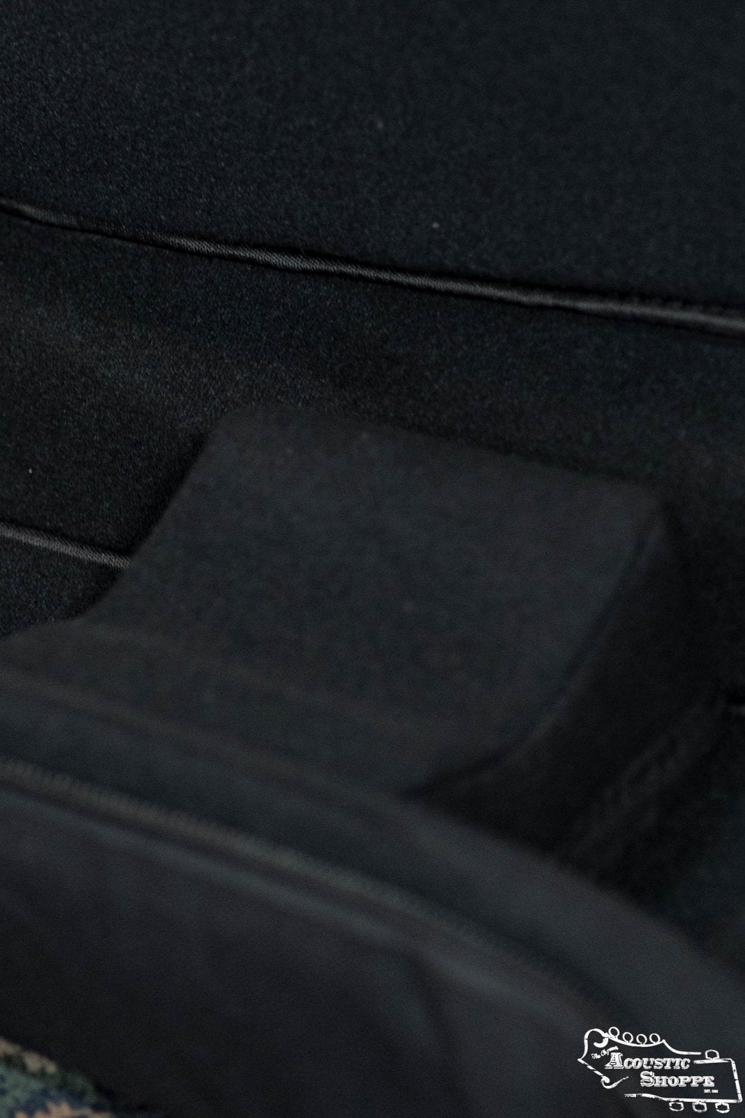 Close-up of the Guild Deluxe Acoustic Gig Bag - Jumbo by Guild Guitars, featuring black fabric, distinct seams, a raised rectangular section with visible stitching, and a small white logo in the lower right corner.