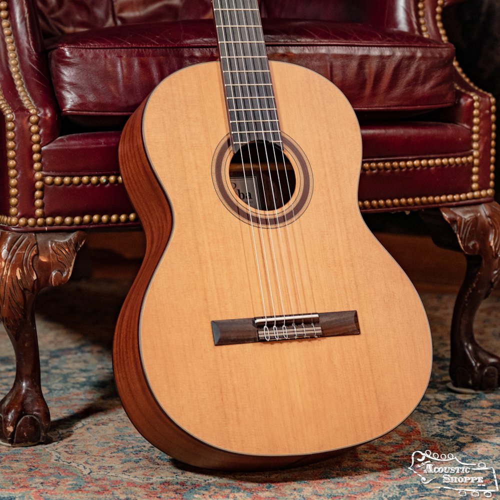The Cordoba C3M Red Cedar/Mahogany Classical Guitar (#8691) by Cordoba stands upright against a red leather chair with brass studs, set on a patterned rug.