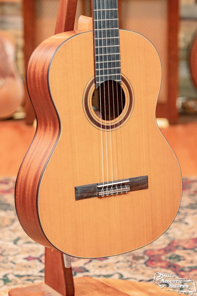 The Cordoba C3M Red Cedar/Mahogany Classical Guitar #8691 is showcased indoors on a stand. It features a solid cedar top and natural wood finish, with other guitars and a patterned rug in the background.