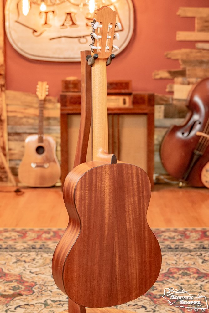 The Cordoba C3M Red Cedar/Mahogany Classical Guitar (#8691) is shown on a stand with its back visible. Nearby are another guitar, a double bass, vintage amp, rustic decor, and a patterned rug under warm lighting.
