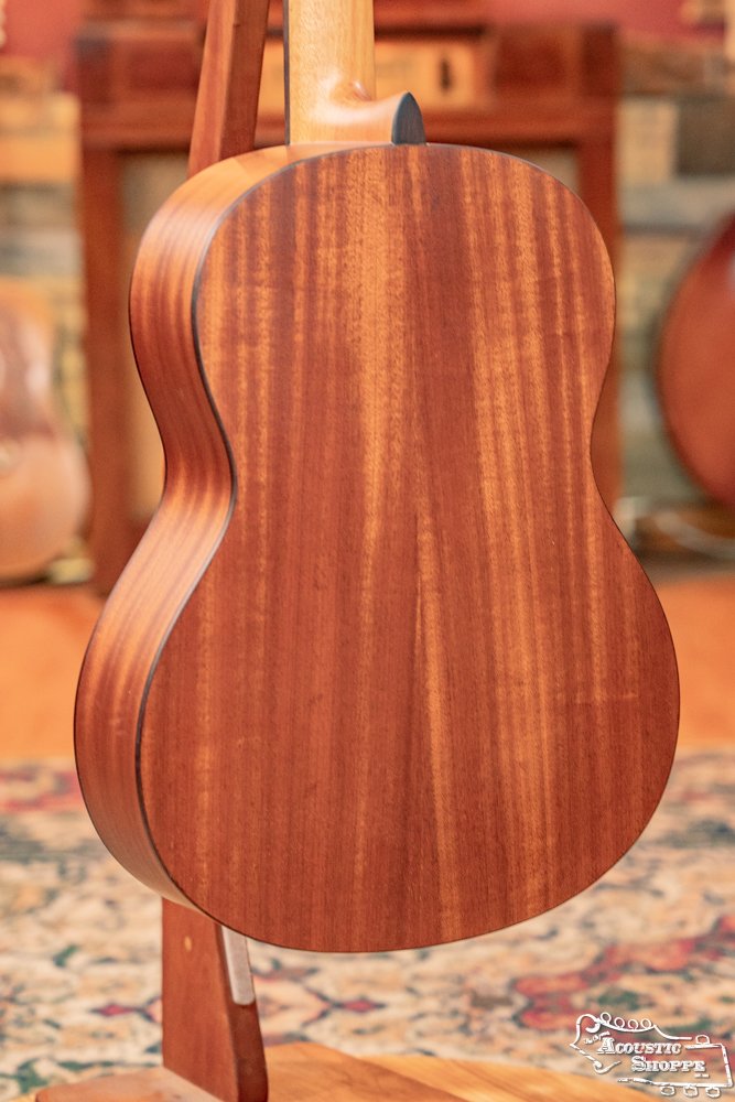 A close-up of the Cordoba C3M Red Cedar/Mahogany Classical Guitar #8691 highlights its smooth wooden back, displayed on a stand with a softly blurred room and patterned rug in the background.