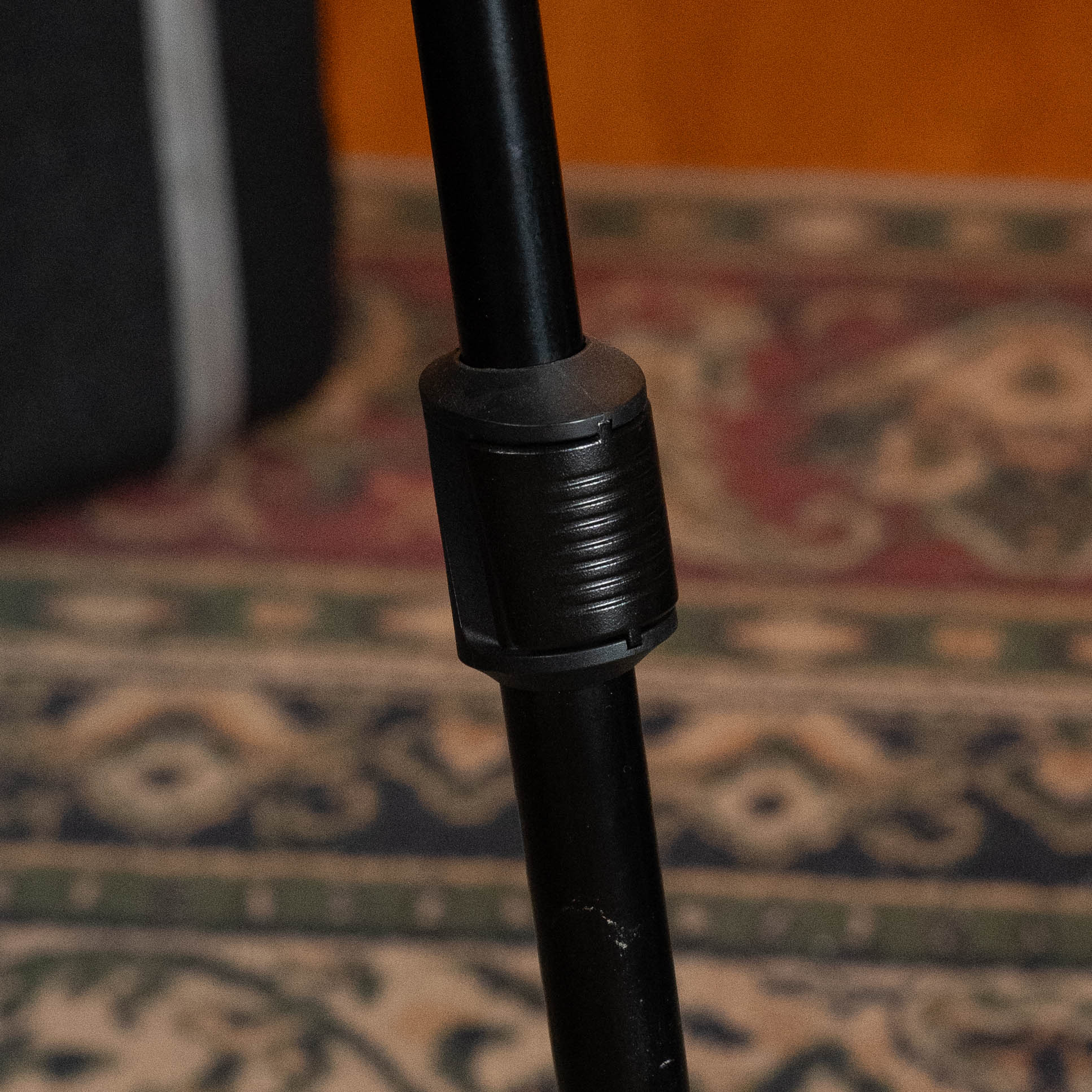 Close-up of a black adjustable tripod leg with locking mechanism, like that on the Hercules GS415B Stand with Auto Grip System by Hercules, seen on a green, red, and beige patterned rug. A dark fabric object appears in the background.