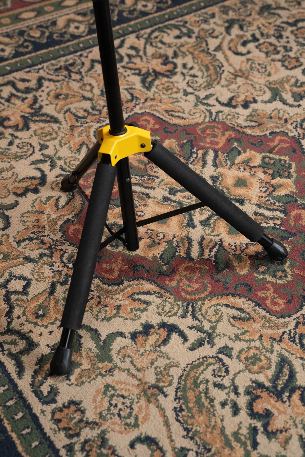 A Hercules Auto Grip Single Guitar Stand w/Foldable Yoke (GS415BPLUS) by Hercules, featuring a yellow joint, is set up on a patterned carpet with beige, green, and red designs.