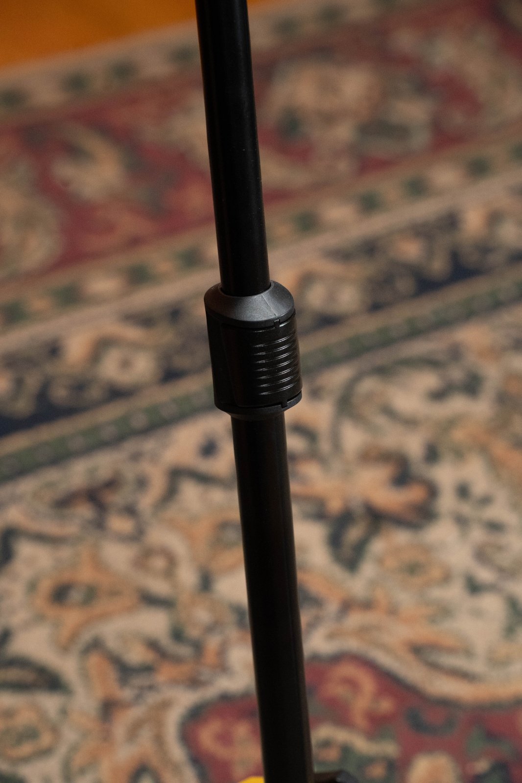 A close-up of the Hercules Auto Grip Single Guitar Stand w/Foldable Yoke (GS415BPLUS) by Hercules, showing its black metal pole with adjustable connector and Auto Grip System, set against a rug with intricate red, blue, and beige patterns.