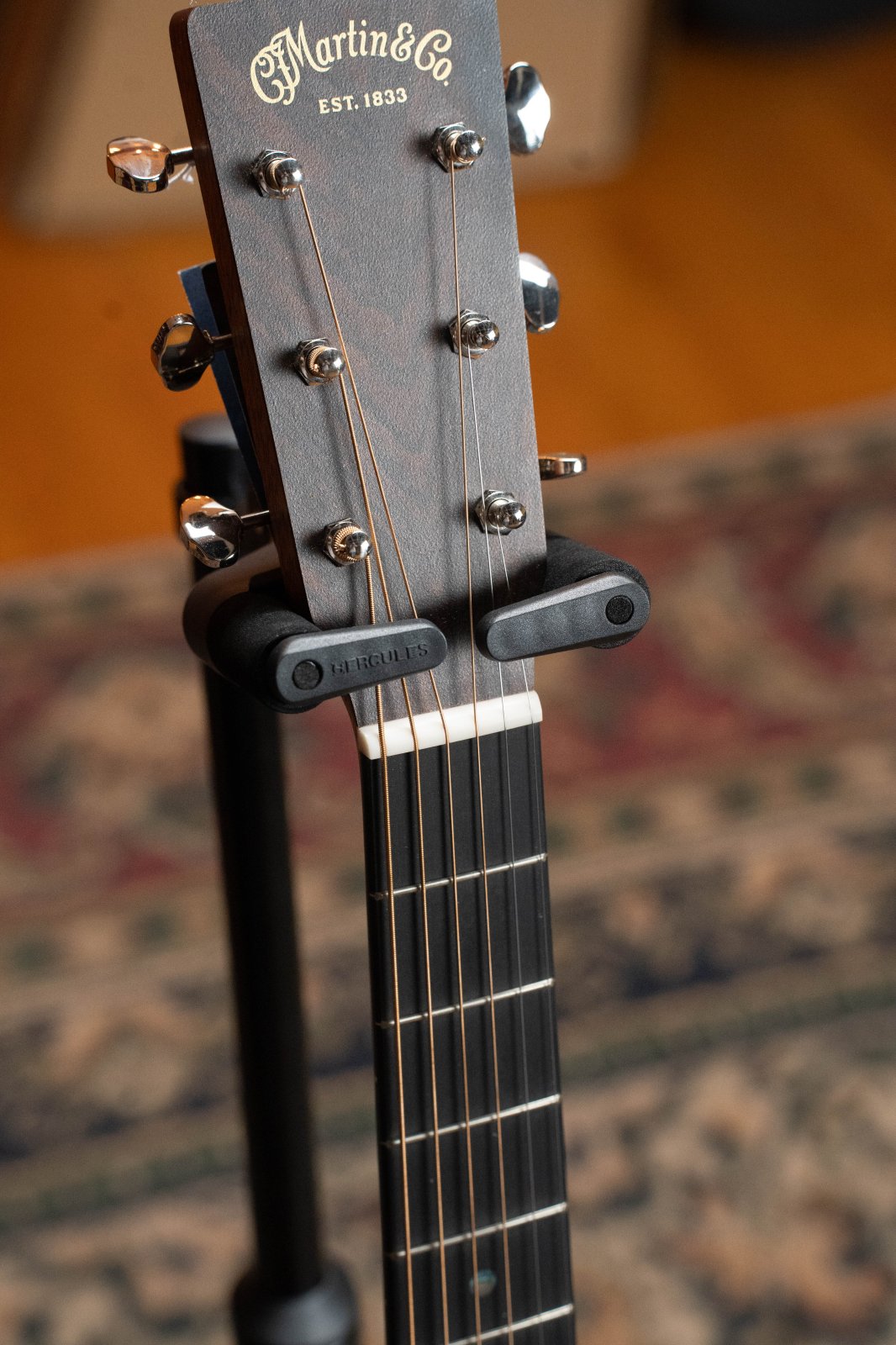 Close-up of a Martin & Co acoustic guitar headstock resting on a Hercules GS415BPLUS Auto Grip Single Guitar Stand with foldable yoke, tuning pegs visible, and a patterned rug softly blurred in the background.