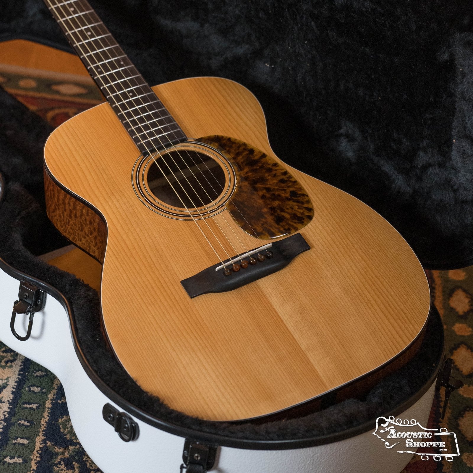 A close-up of a Hinde Adirondack/Quilted Sapele OO 14-Fret Acoustic Guitar #6 with a quilted Sapele finish, dark pickguard, and six strings rests in an open white plush-lined hard case on a patterned rug.